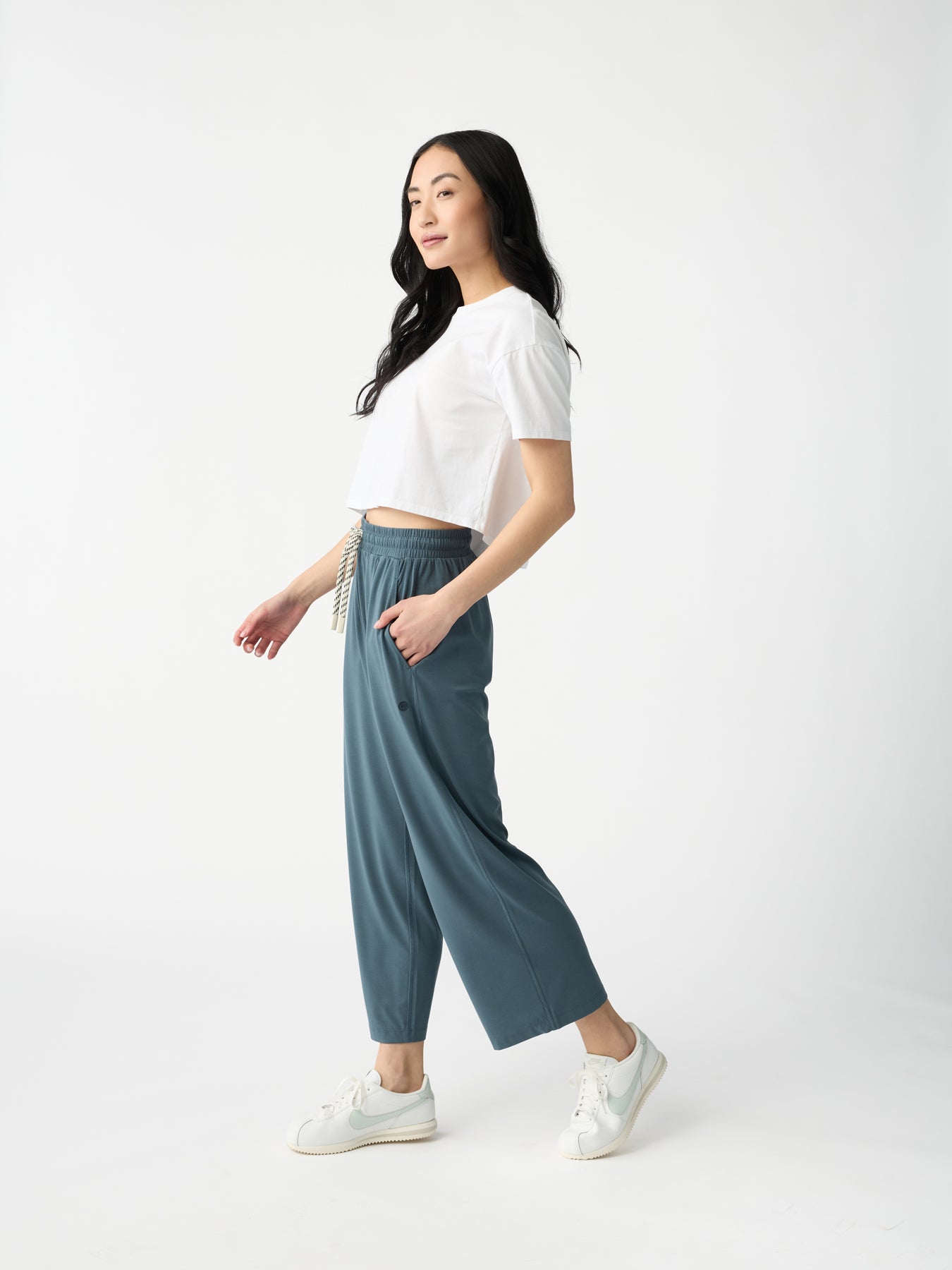 A person with long black hair in a white short-sleeve shirt, Cozy Earth's Women's Studio Cropped Wide Leg Pant, and white sneakers walks left against a plain white background. 