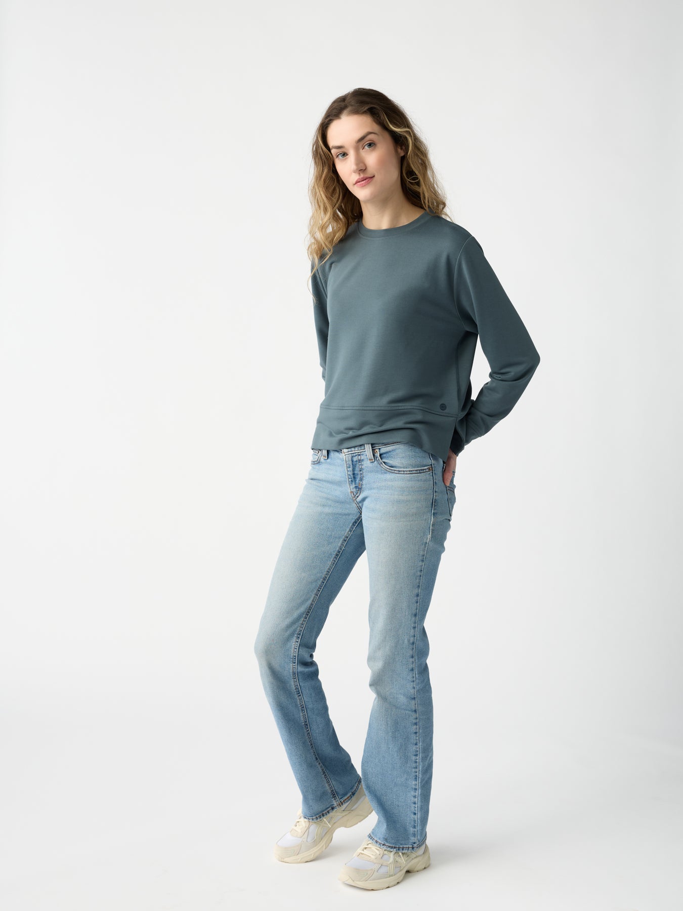 A person with long hair leans casually against a plain background, wearing Cozy Earth's Women's StudioLite Crewneck in blue-green, light blue jeans, and sneakers, with their hands tucked in their jeans' pockets. 