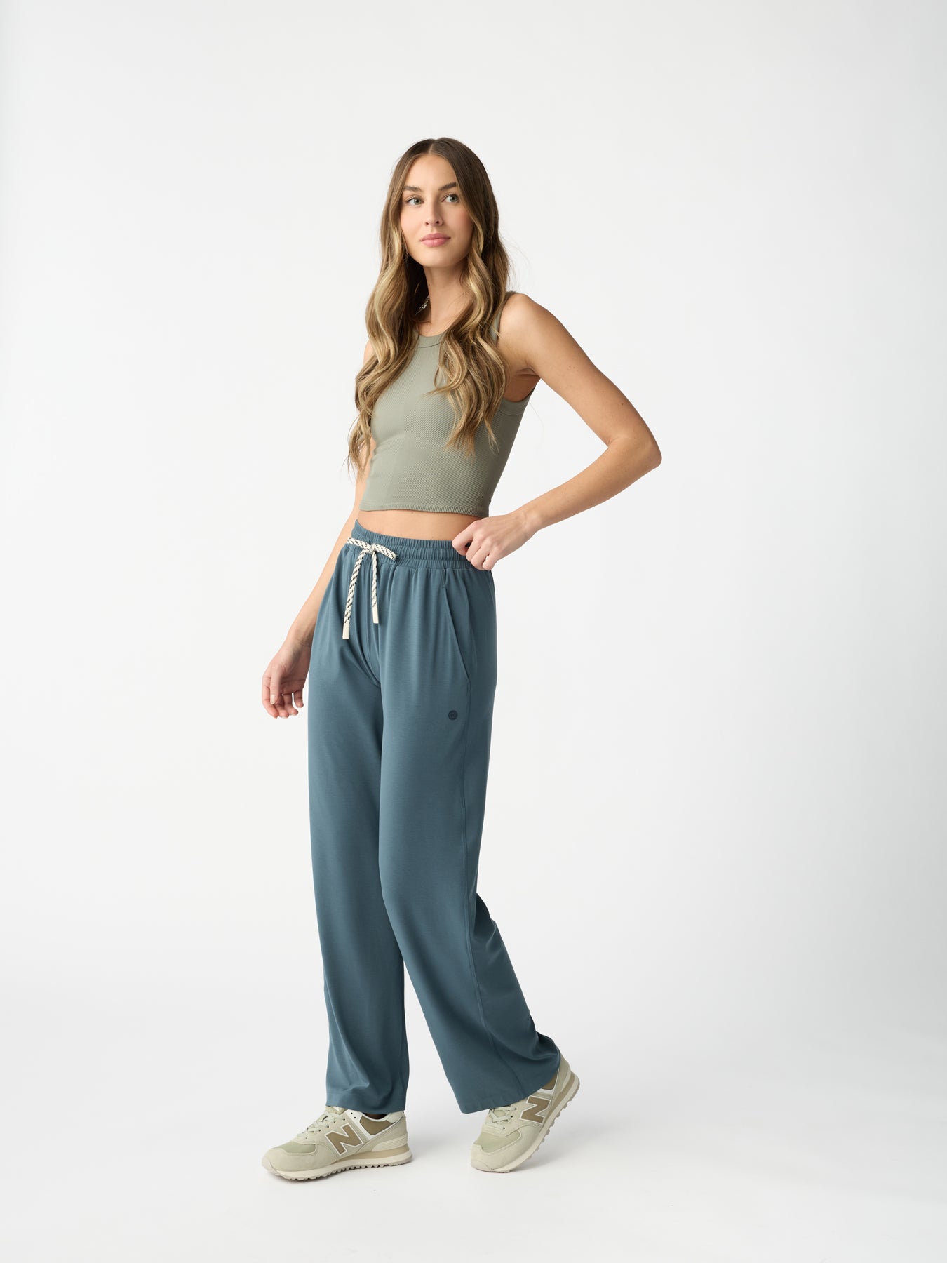 A person with long hair in a sleeveless green top and Cozy Earth's blue Studio Wide Leg Pants stands with one hand in pocket, wearing green sneakers against a white background. |Color:Dawn