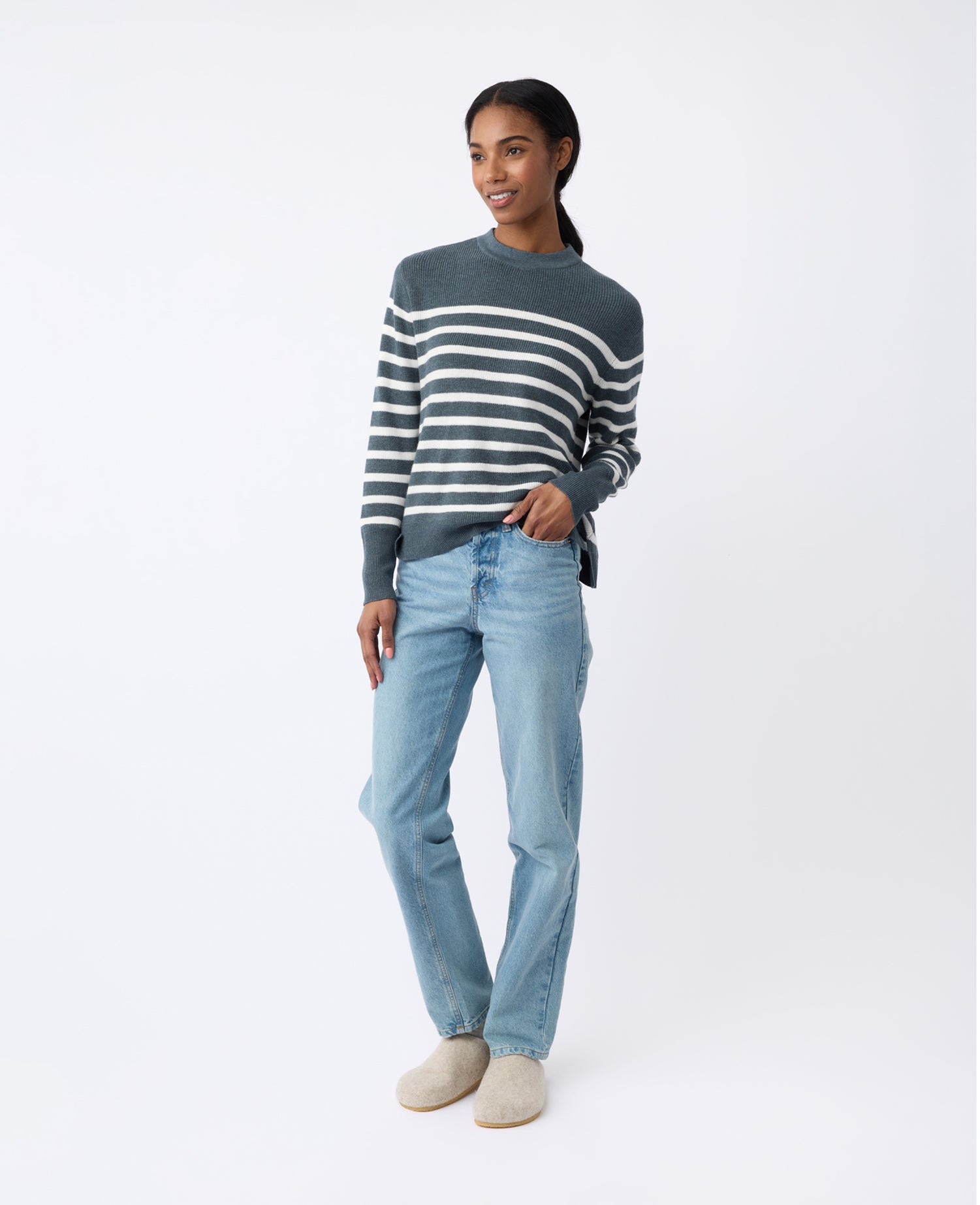 A person smiles while standing against a plain background, dressed in a Women's Rowan Sweater from Cozy Earth, paired with light blue jeans and beige shoes. They pose casually with one hand in their jeans pocket. 