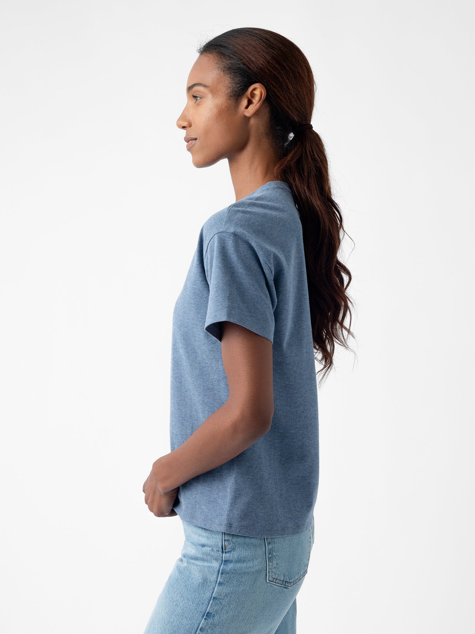 A person with long, wavy brown hair tied back in a ponytail stands in profile, facing left, wearing Cozy Earth's Women's All Day Tee in blue and light blue jeans. The background is plain white. 