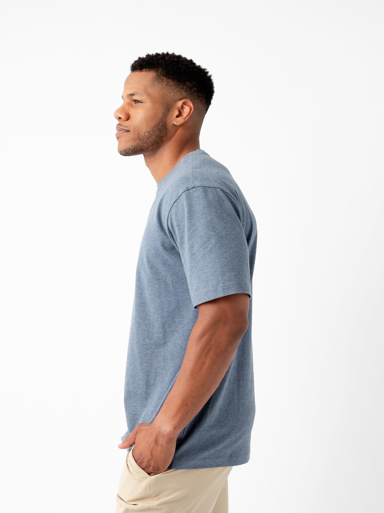 A man wearing Cozy Earth's Men's All Day Tee in a blue short-sleeve design and beige pants is standing in profile against a white background. He has short hair and a trimmed beard, looking straight ahead with a neutral expression. 