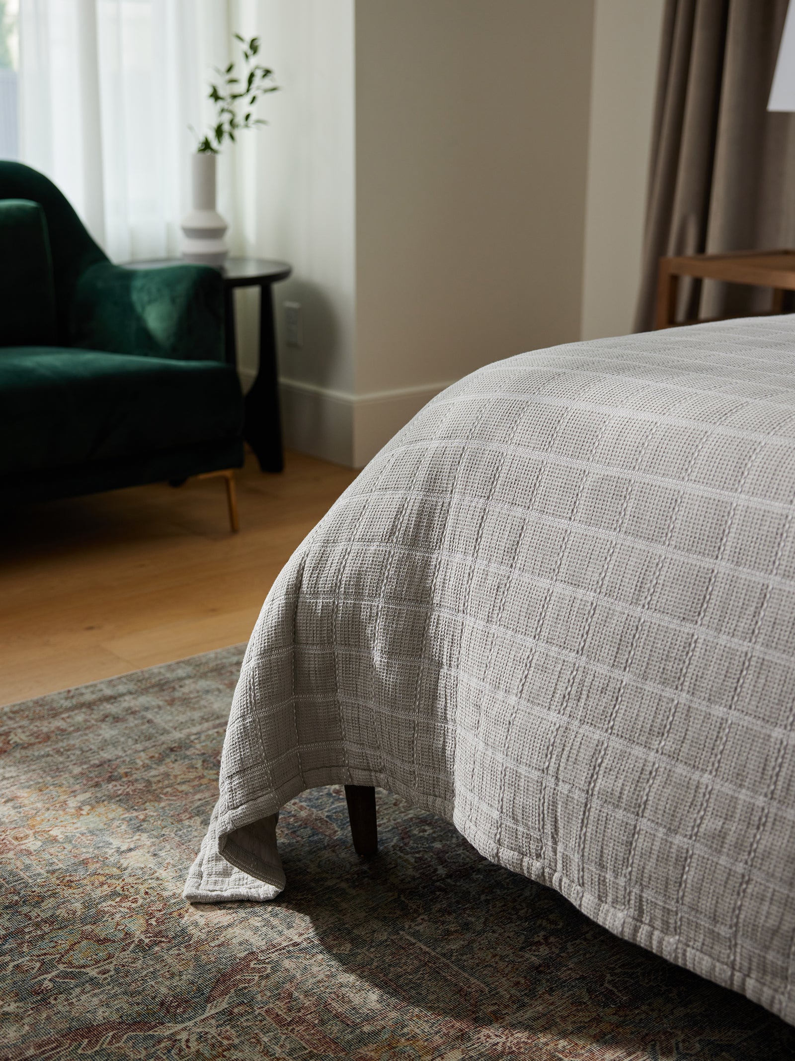 A cozy bedroom corner with sunlight streaming through a window. The bed is adorned with a neatly made Waffle Windowpane Blanket from Cozy Earth. A dark green velvet chair sits beside a small round table with a plant. The floor features a patterned rug and light wooden flooring. 