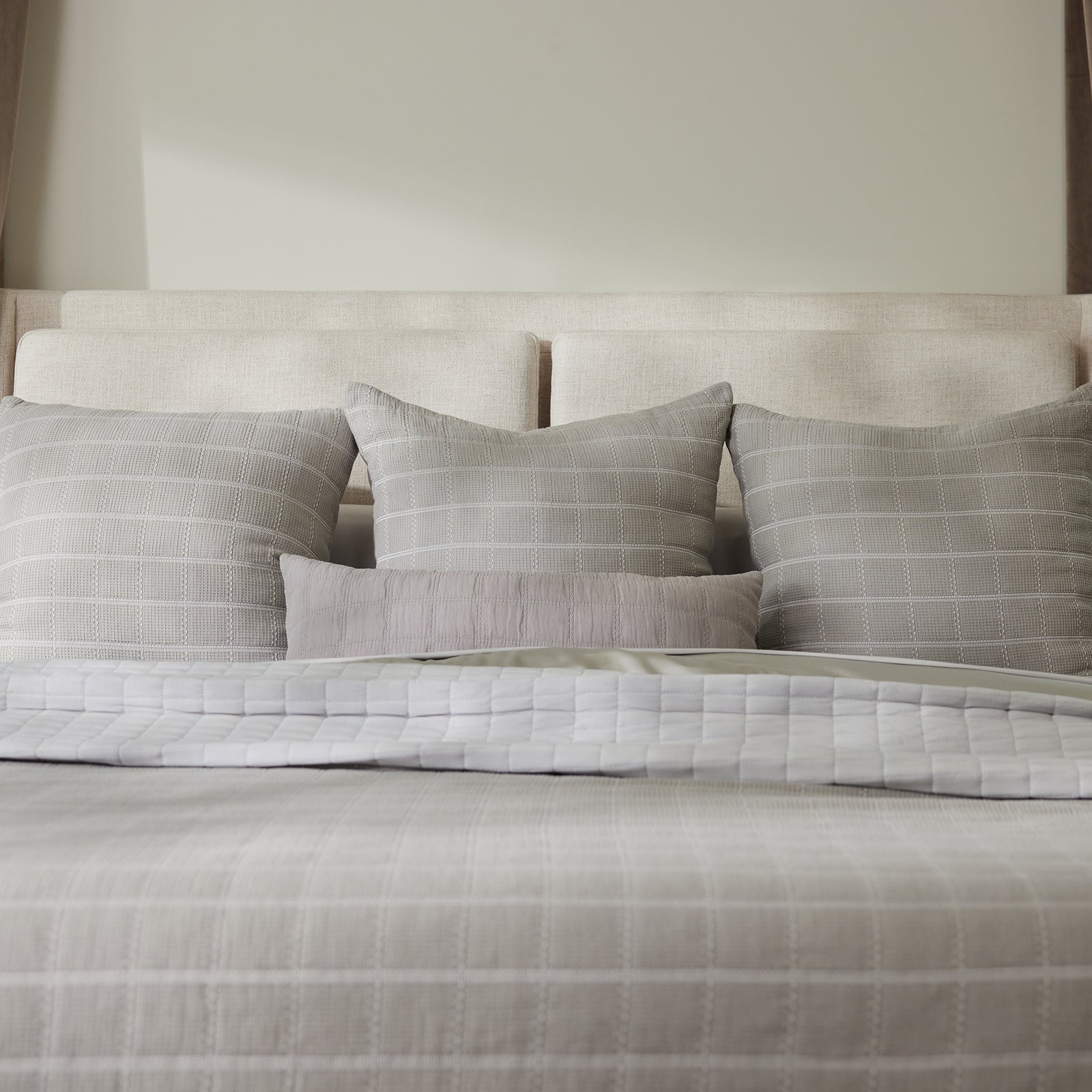 A neatly made bed featuring a beige upholstered headboard. The bed is dressed in a light gray duvet cover with a checked pattern and adorned with five pillows: three larger Waffle Windowpane Pillows by Cozy Earth at the back, along with two smaller accent pillows—one solid gray and one striped. 