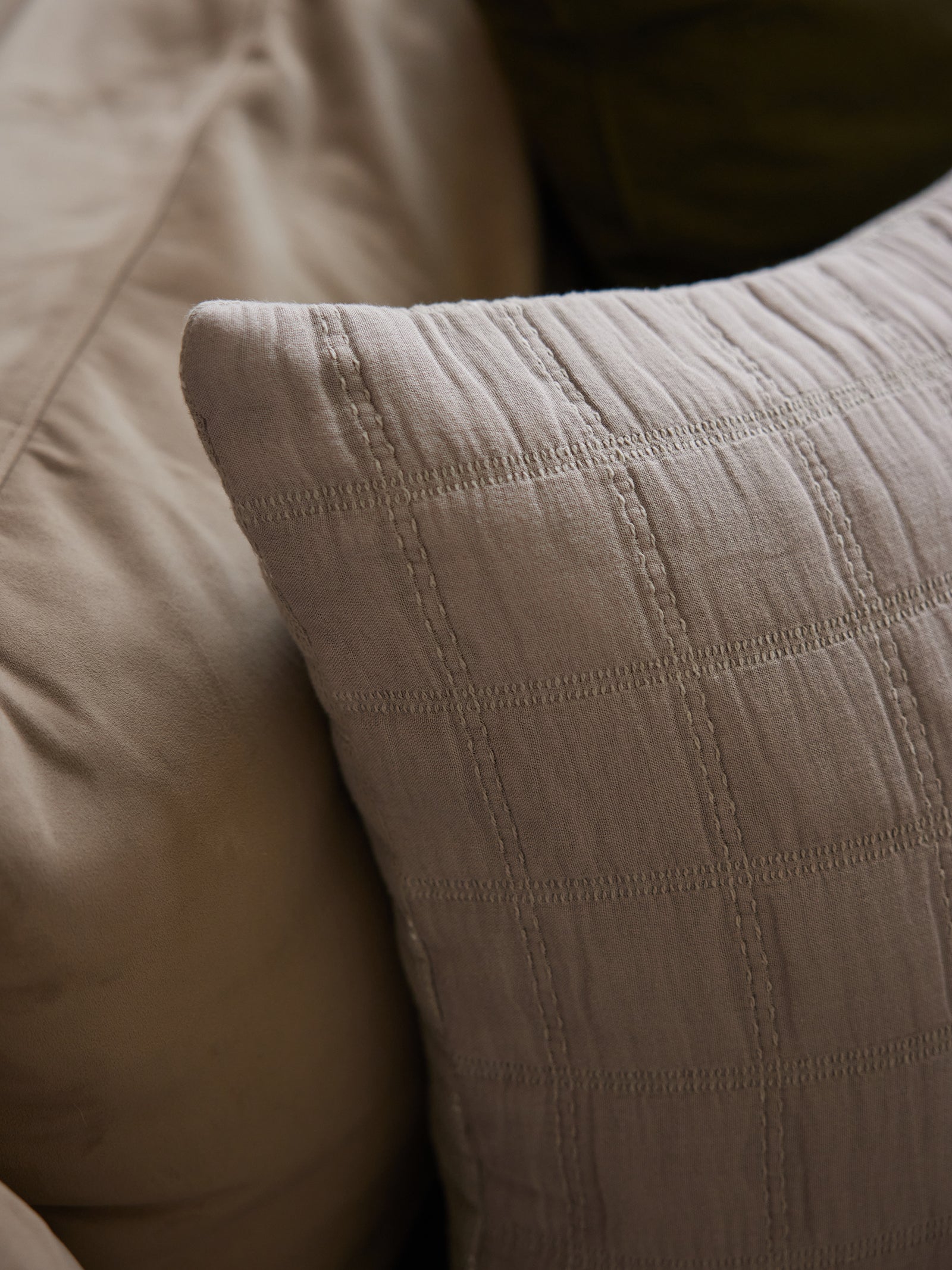 A close-up image of the Waffle Windowpane Pillow by Cozy Earth in light grey, featuring a textured grid pattern. The pillow is placed against what appears to be a neutral-colored sofa or bedspread. The soft texture and simple design give a cozy and elegant appearance. 