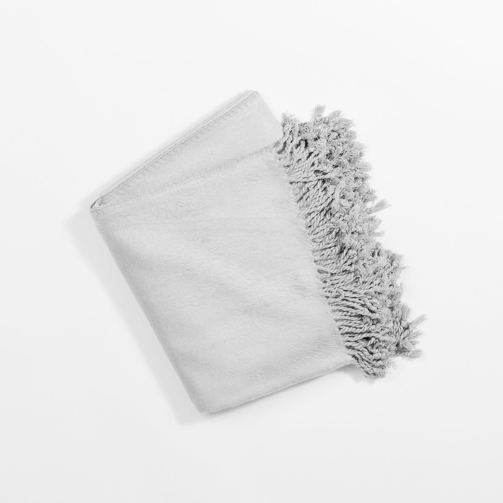 A folded Bamboo Tassel Throw from Cozy Earth, in a light gray color, lies on a white background. The throw appears soft and plush, with wavy and textured fringes that add a decorative touch to its otherwise simple design. 