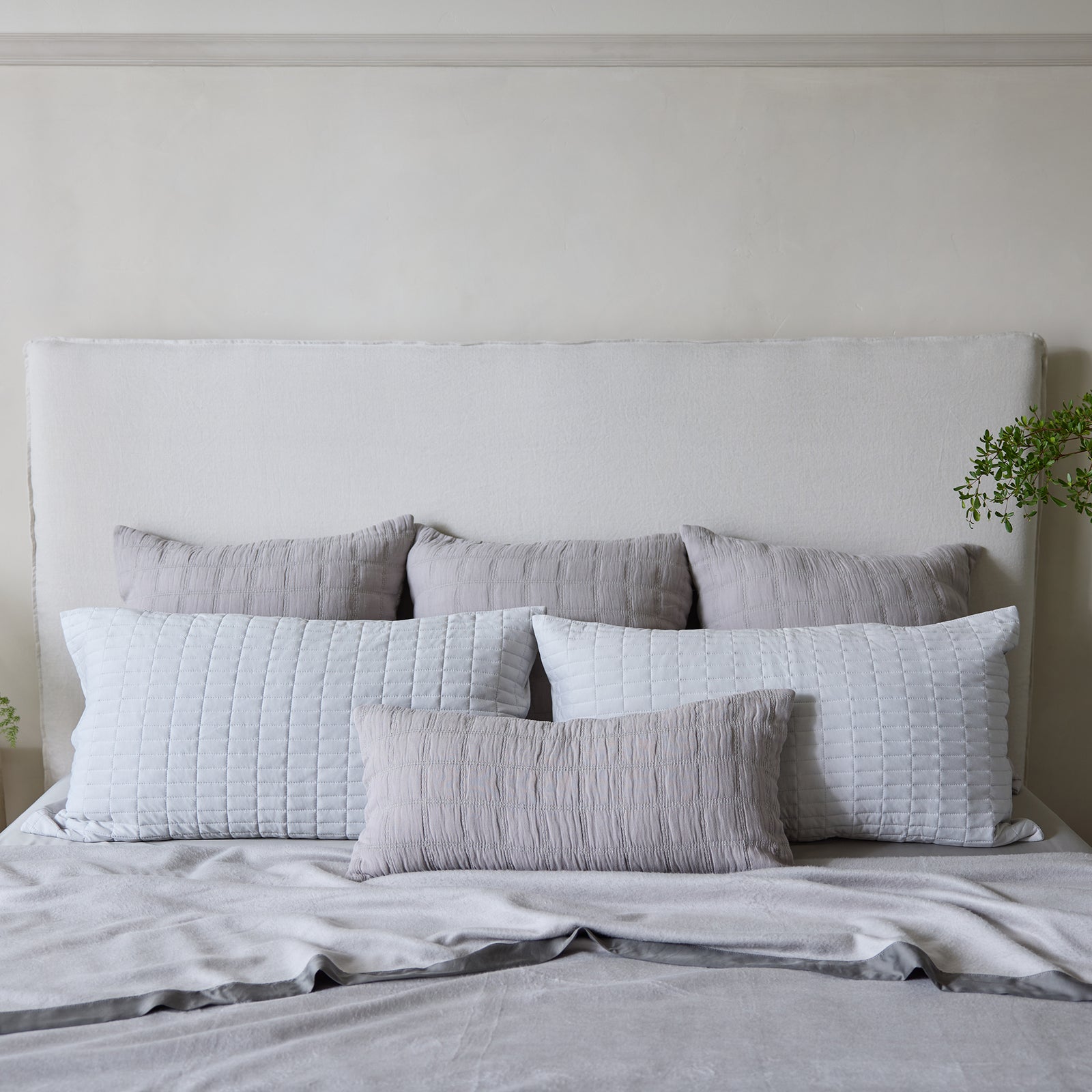 A neatly made bed with a white quilted headboard features light gray and white pillows arranged in a row, topped with the luxurious Bamboo Blanket from Cozy Earth. The set of pillows includes three larger white pillows at the back, three smaller gray pillows in front, and a long gray rectangular pillow in front. Minimalistic decor with some green plants visible on either side enhances the calming ambiance. 