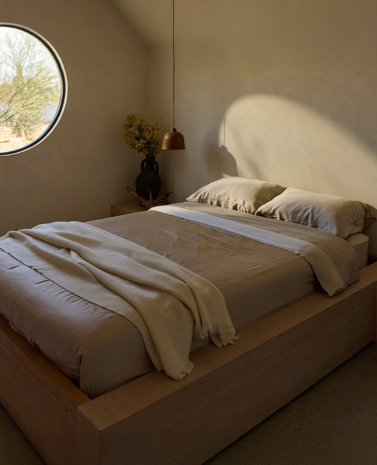 A minimalist bedroom features a low wooden platform bed with the Bamboo Bedding Deluxe Bundle by Cozy Earth in beige, and a cozy blanket. Soft light streams through a round window. A side table holds a dark vase with yellow flowers under a modern pendant lamp, all in neutral tones. 