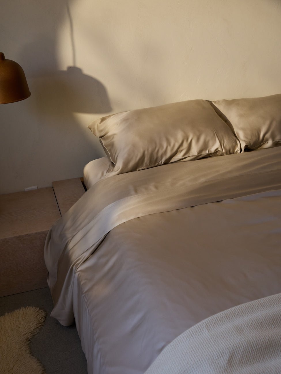 A neatly made bed with Cozy Earth's Bamboo Fitted Sheet and pillows is in a softly lit room. A wooden side table is next to the bed. A copper-colored lamp hangs overhead, casting a shadow on the light-colored wall, and a fluffy rug is partially visible on the floor. 