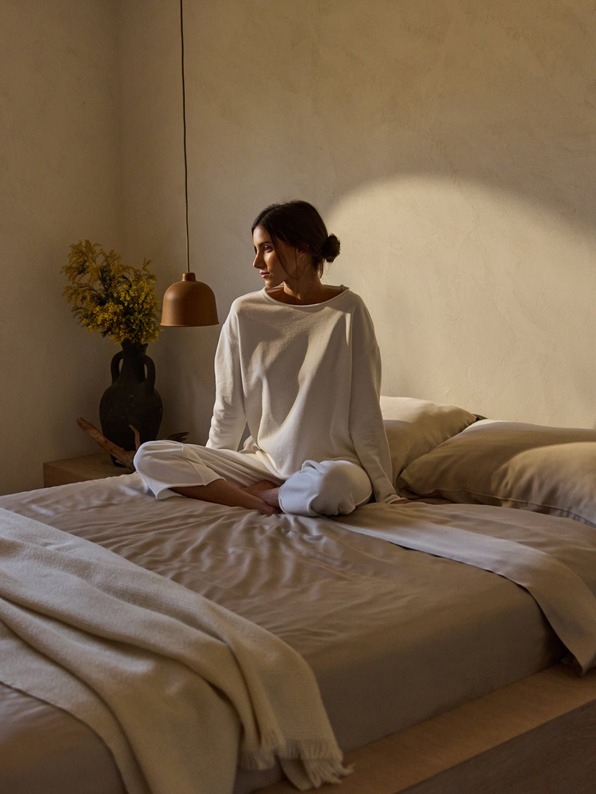 Dressed in white, a person sits cross-legged on a bed from the Cozy Earth Bamboo Bedding Core Bundle. Soft sunlight highlights their face as they gaze out, with neutral-toned decor around them, including a black vase with flowers on the bedside table and a hanging lamp overhead. 