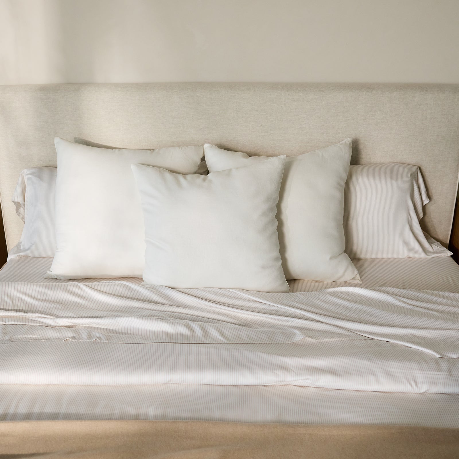 A neatly made bed, featuring a beige fabric headboard, is adorned with four white pillows and topped with the Bamboo Duvet Cover by Cozy Earth. Sunlight softly illuminates the bedding, creating a serene and inviting atmosphere. 