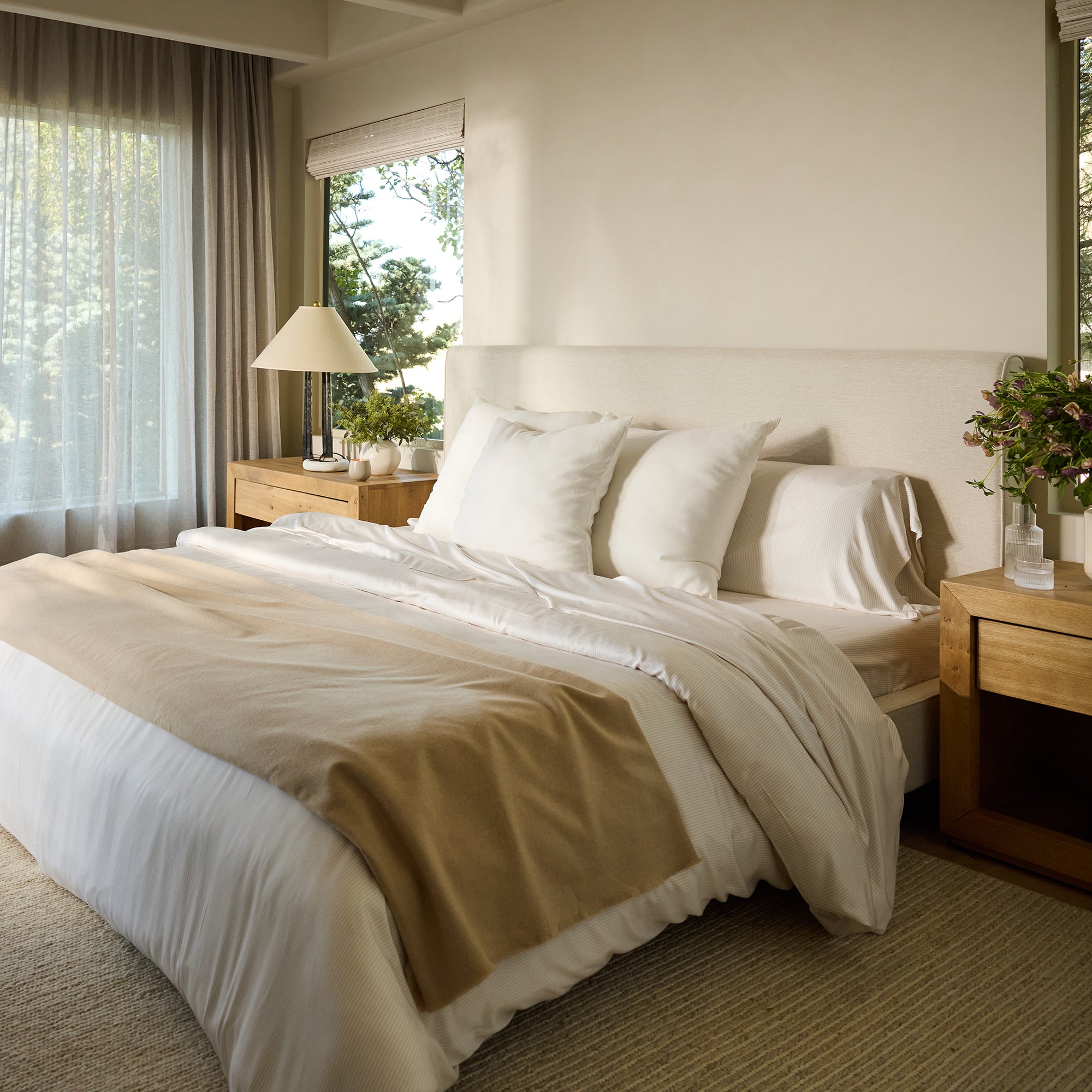 A cozy bedroom with a neatly made bed featuring Cozy Earth's bamboo fitted sheet in white, complemented by a beige blanket. Wooden nightstands flank the bed, each adorned with a lamp, and one displaying a vase of flowers. Sunlight filters through sheer curtains against large windows. 