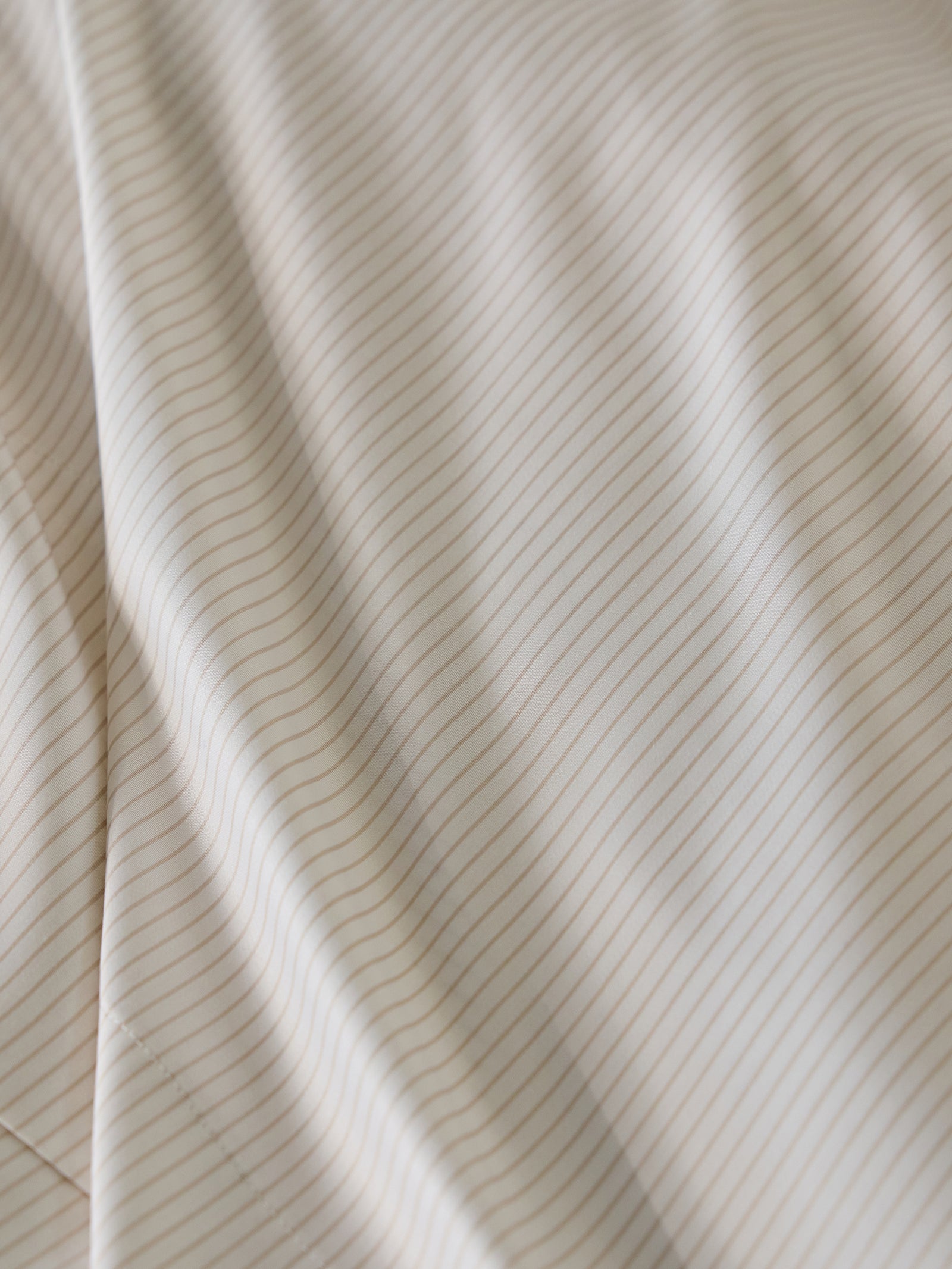 Close-up of the Bamboo Flat Sheet by Cozy Earth, featuring a beige fabric with thin, evenly spaced horizontal lines in a light brown color. The fabric appears to be soft and smooth, with gentle folds and subtle shadows enhancing its texture. 