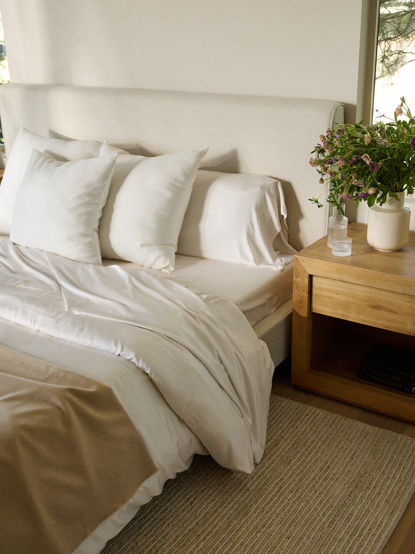 A neatly made bed features the Bamboo Sheet Set by Cozy Earth, complete with two pillows and a light beige blanket. A wooden nightstand holds a vase of purple and green flowers alongside a small clear bottle. Soft light filters through a nearby window. 