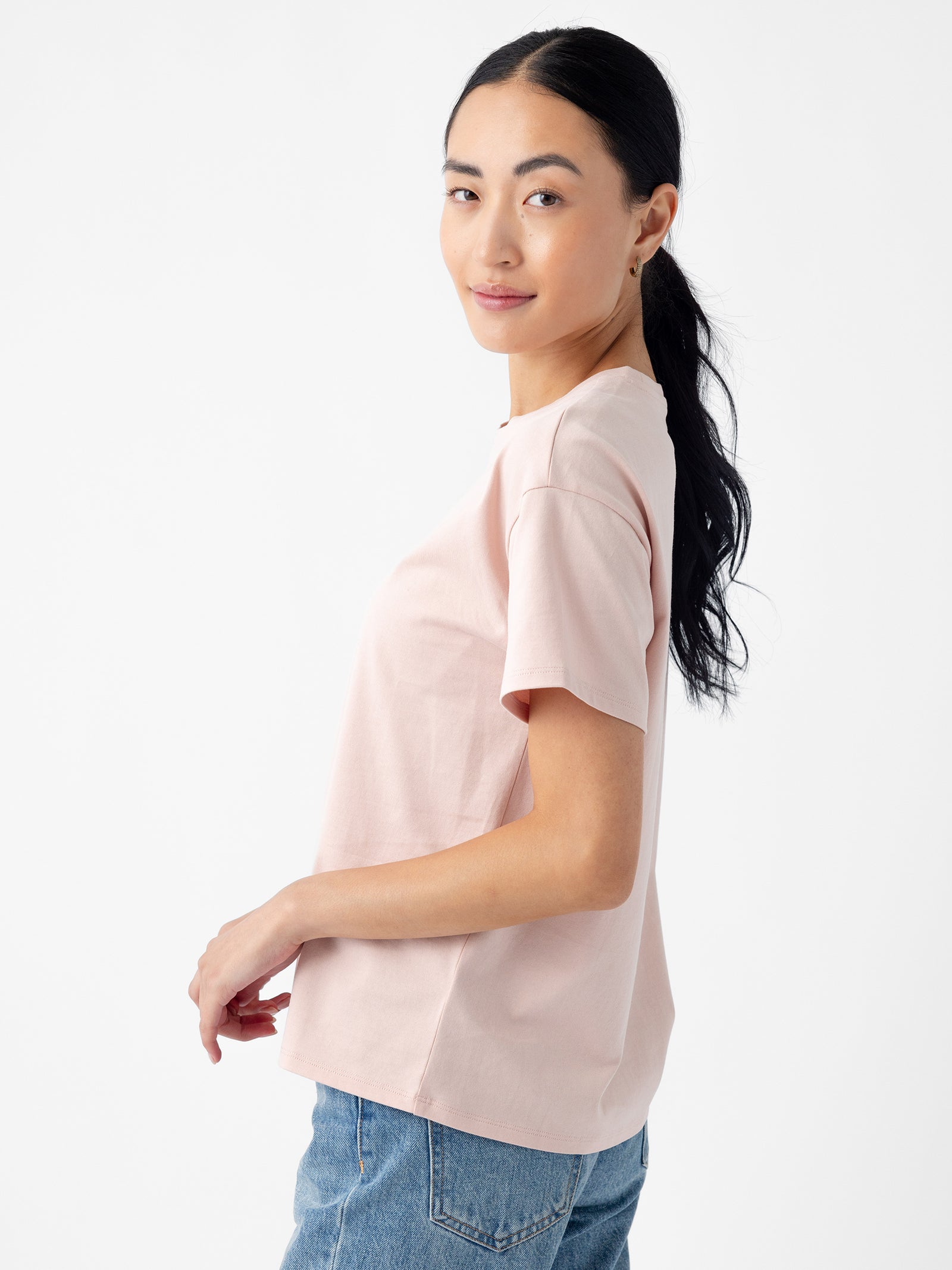 A person with long dark hair is seen from the side, wearing Cozy Earth's Women's All Day Tee in light pink and blue jeans. They are smiling slightly and have their hair tied back in a ponytail. The background is plain white. 