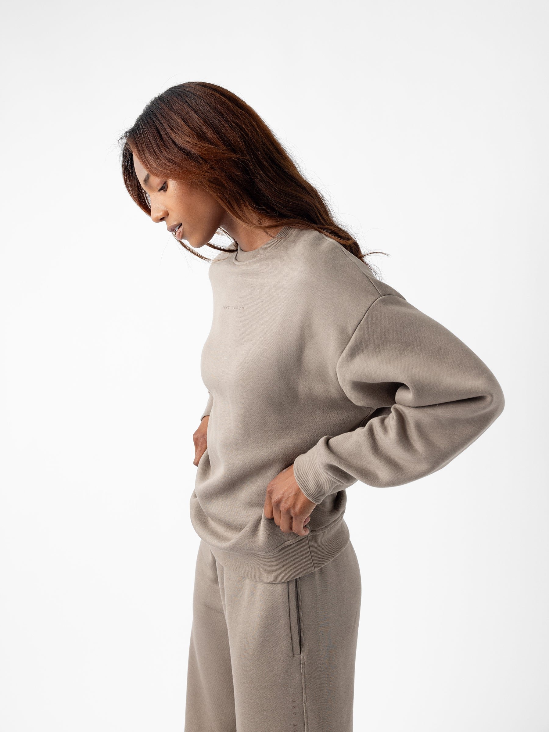 A woman with long, brown hair is standing in profile against a white background. She is wearing the Cozy Earth's Women's CityScape Crewneck sweatshirt and matching pants in light brown, with her hands resting on her hips and looking downward. The outfit appears comfortable and casual. 