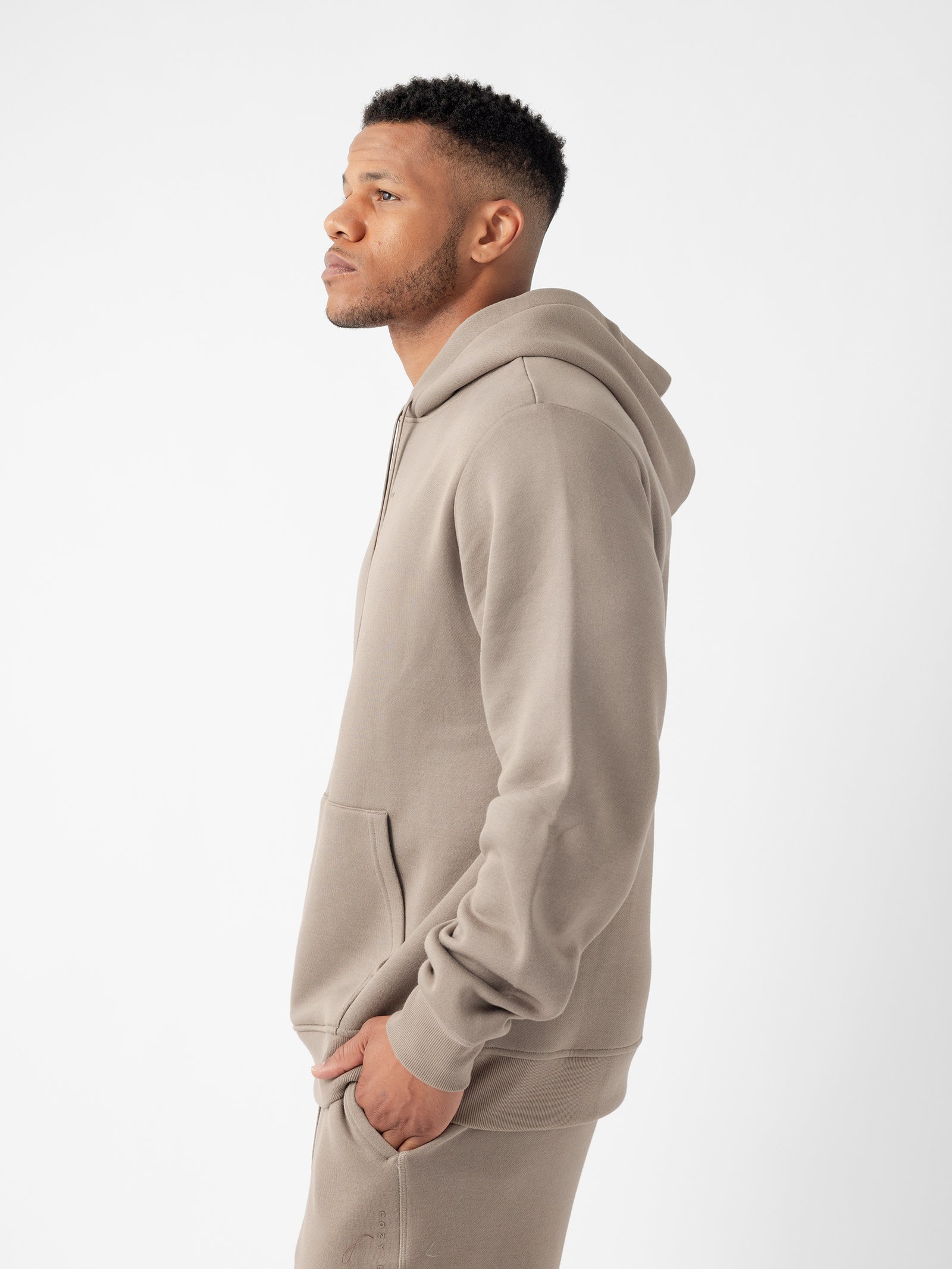 A man stands in profile against a white background, wearing a light brown Men's CityScape Hoodie from Cozy Earth. His hands are in the front pocket of the hoodie, and he gazes to the left with a relaxed expression. The hoodie has a simple, clean design without any visible logos or text. 