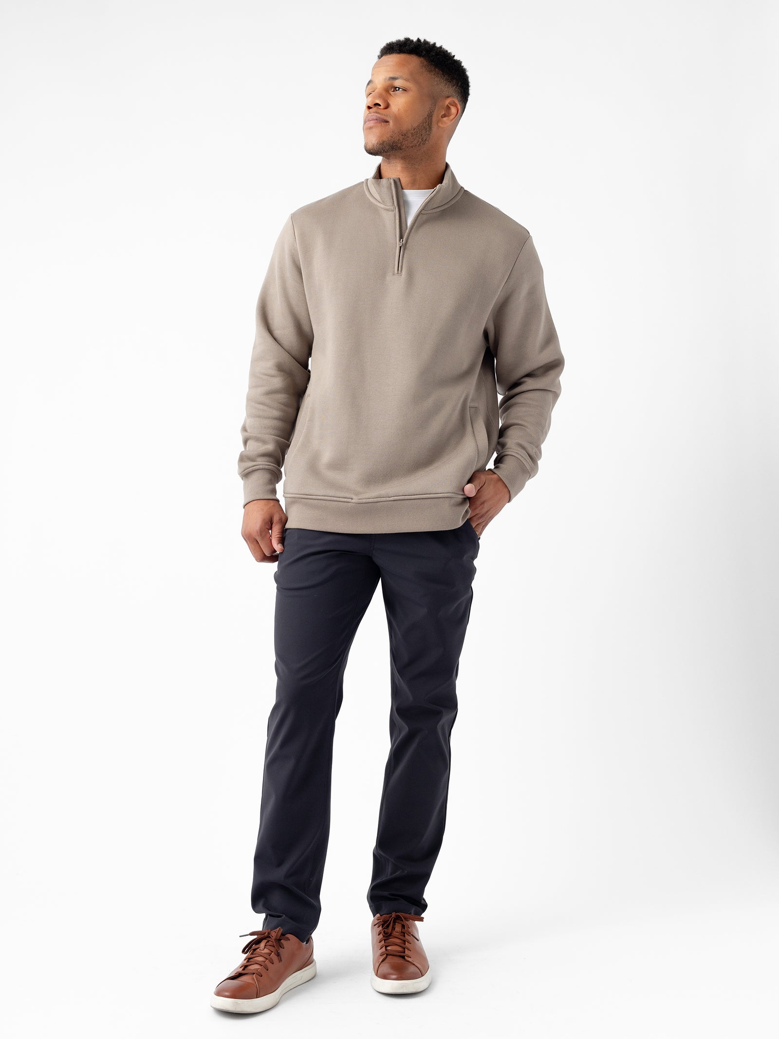 A man stands against a plain white background, gazing upward to his right. He is wearing Cozy Earth's Men's CityScape Quarter Zip in taupe over a white shirt, paired with black pants and brown leather sneakers. His hands are in his pockets, exuding a calm expression. 