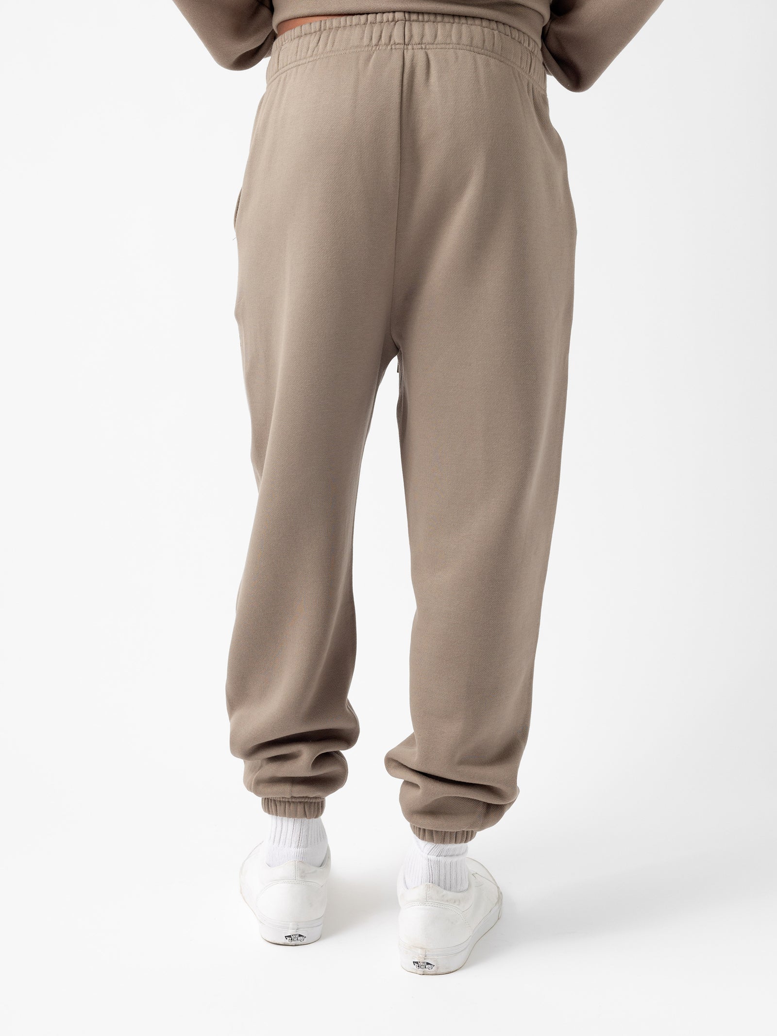 The image shows a person from the rear, wearing Cozy Earth's Men's CityScape Sweatpant in beige, featuring a drawstring waist and elastic cuffs. The pants have a relaxed fit, and the individual is standing upright. The person is also wearing white sneakers and white socks against a plain white background. 