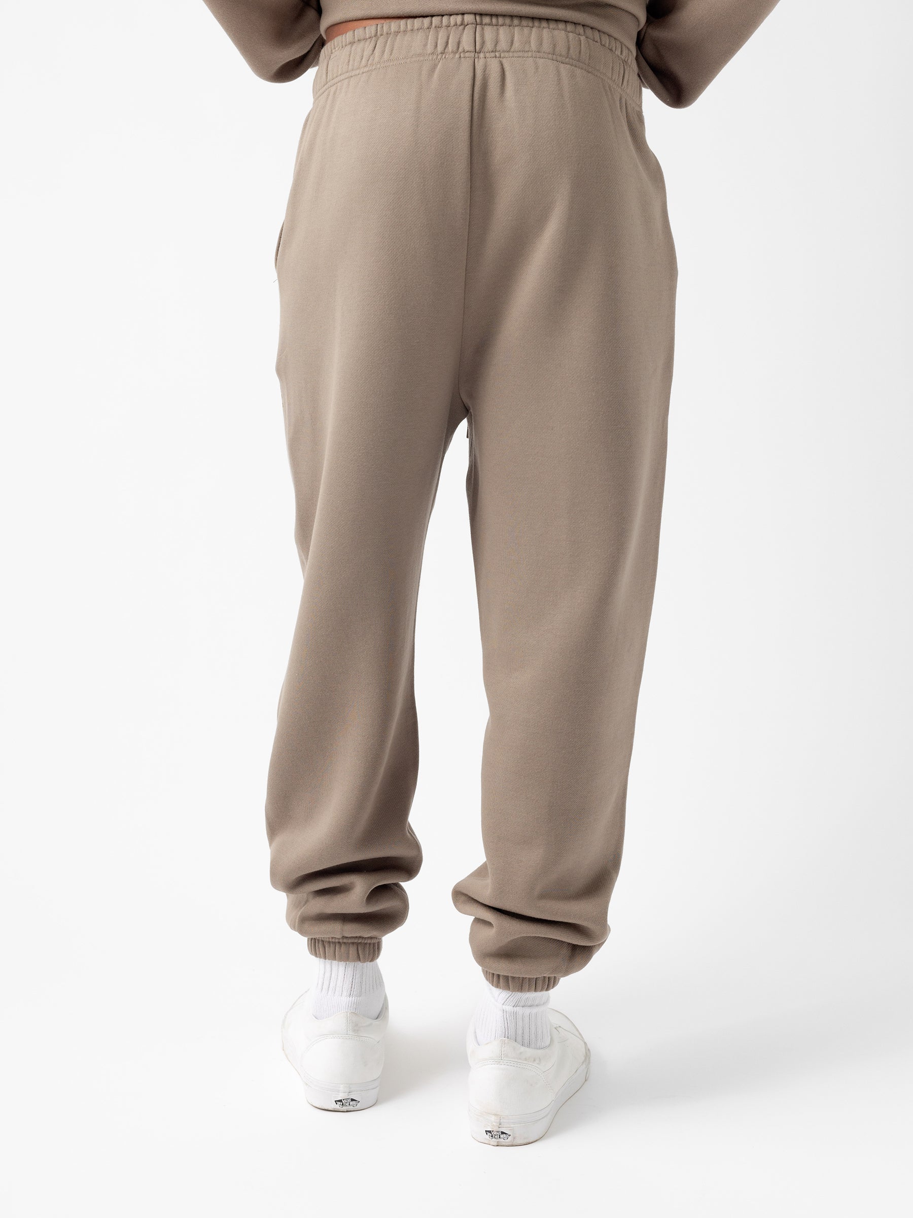 The image shows a person from the rear, wearing Cozy Earth's Men's CityScape Sweatpant in beige, featuring a drawstring waist and elastic cuffs. The pants have a relaxed fit, and the individual is standing upright. The person is also wearing white sneakers and white socks against a plain white background. |Color:Ebony