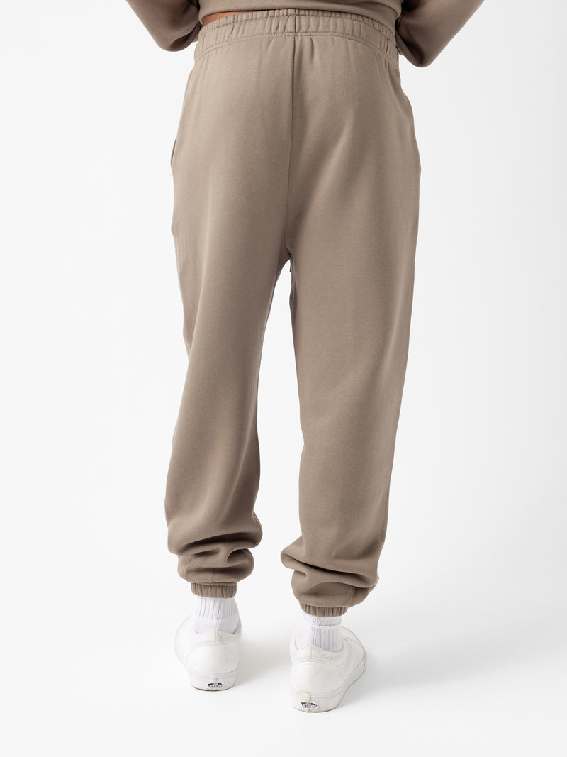 The image shows a person from the rear, wearing Cozy Earth's Men's CityScape Sweatpant in beige, featuring a drawstring waist and elastic cuffs. The pants have a relaxed fit, and the individual is standing upright. The person is also wearing white sneakers and white socks against a plain white background.
