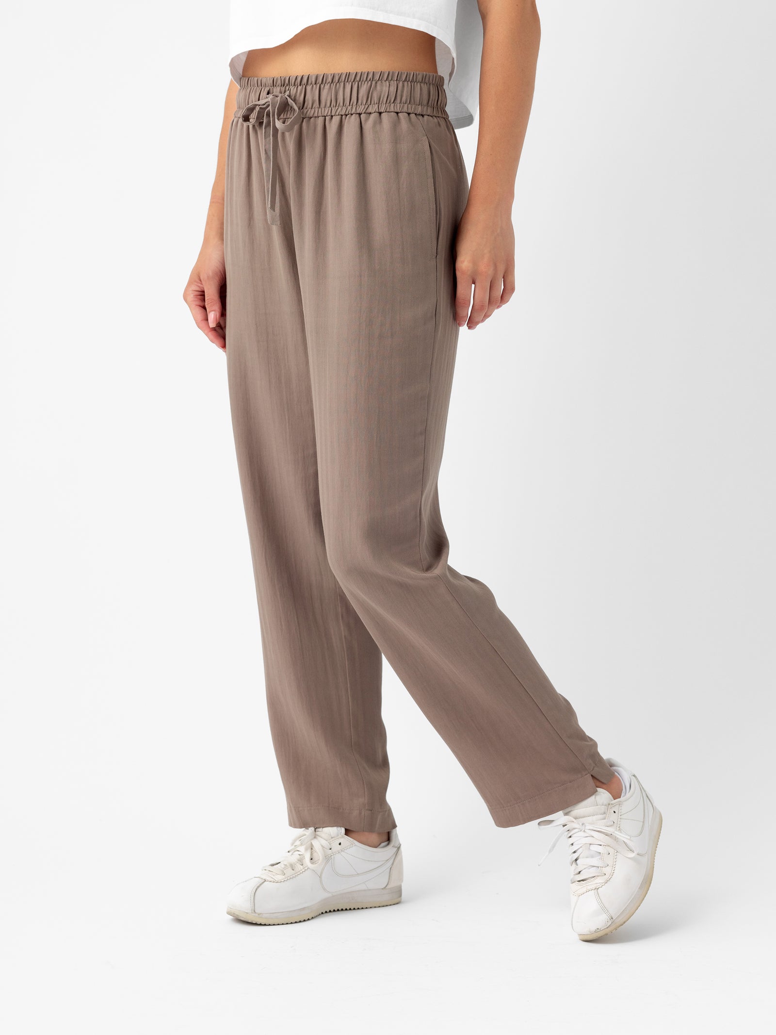 A person is standing against a plain white background, wearing Cozy Earth's Women's Sunset Cropped Pant in taupe. The loose-fitting pants feature a drawstring waist and are paired with white sneakers and a partial view of a white top. The scene is captured from the mid-torso down. 