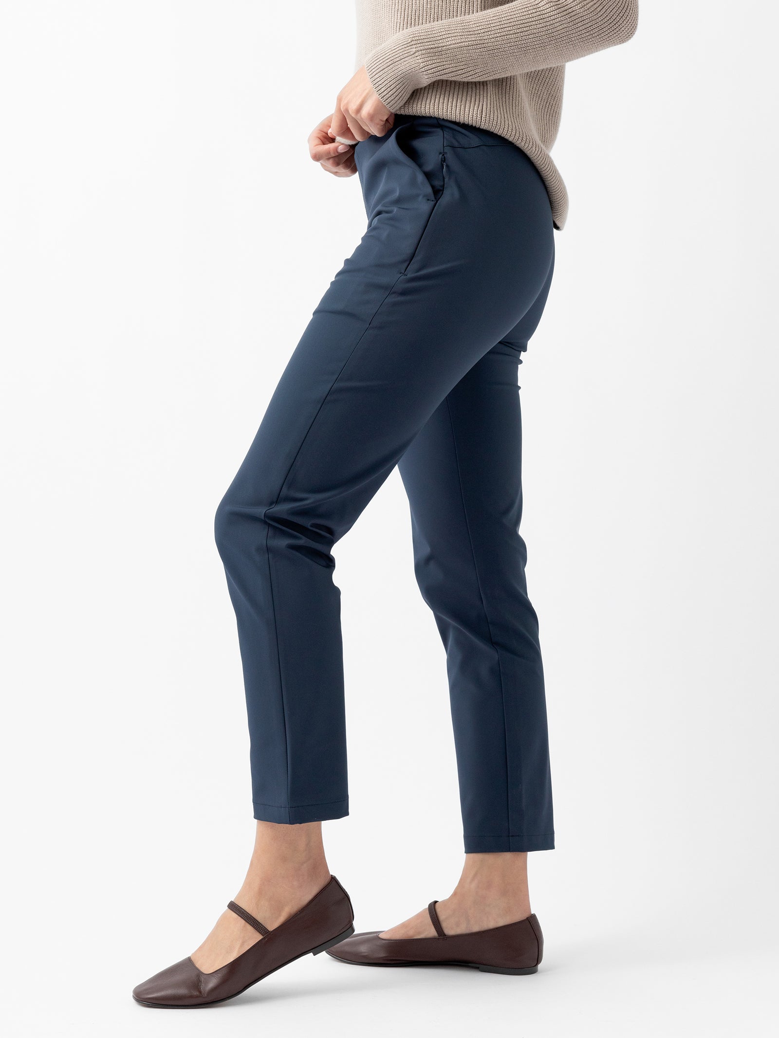 A person is shown from the side, modeling navy blue Women's Always Cropped Pant by Cozy Earth, featuring a tailored fit. They are also wearing a beige sweater and brown flats. The plain white background emphasizes the clothing. 