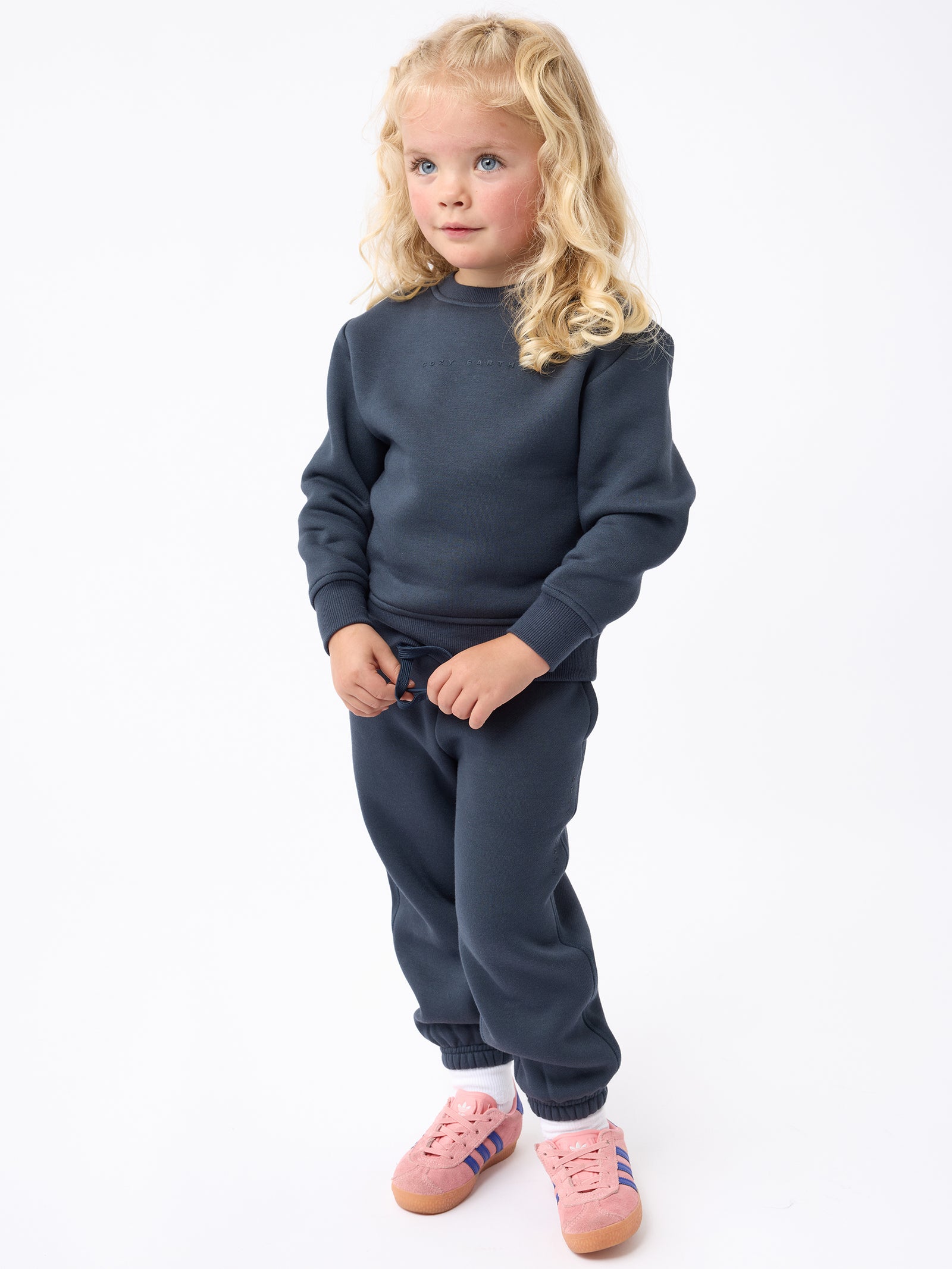 A young child with long blonde hair stands against a white background, wearing a Cozy Earth Kid's CityScape Crewneck and matching dark blue sweatpants. They sport pink sneakers accented with blue and white stripes and look thoughtfully to the side. 