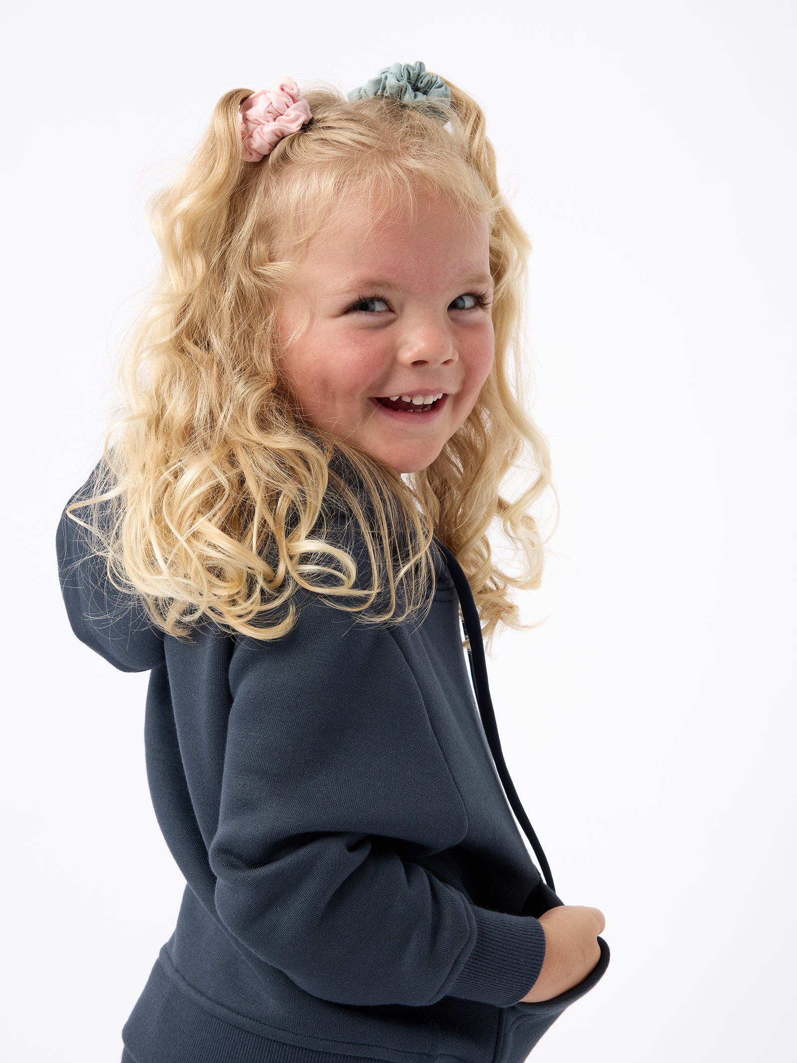 A young girl with blonde curly hair beams as she wears a Kid's CityScape Hoodie by Cozy Earth. Her hair is styled with pink and blue scrunchies against a plain white background. 