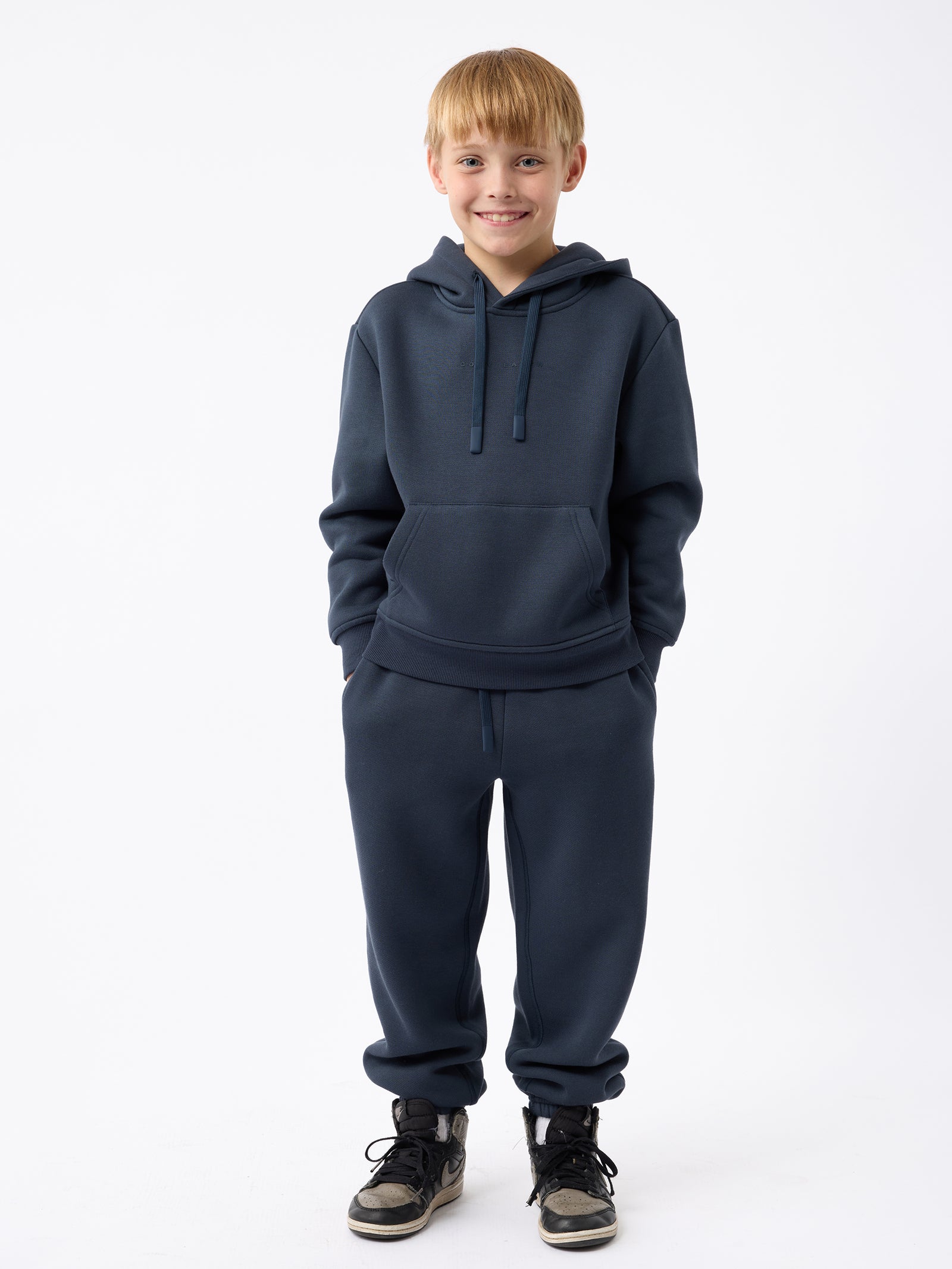 A smiling young boy stands facing the camera, wearing Cozy Earth's Kid's CityScape Sweatpants with a dark blue hoodie. He has his hands in his pockets against a plain white background. 