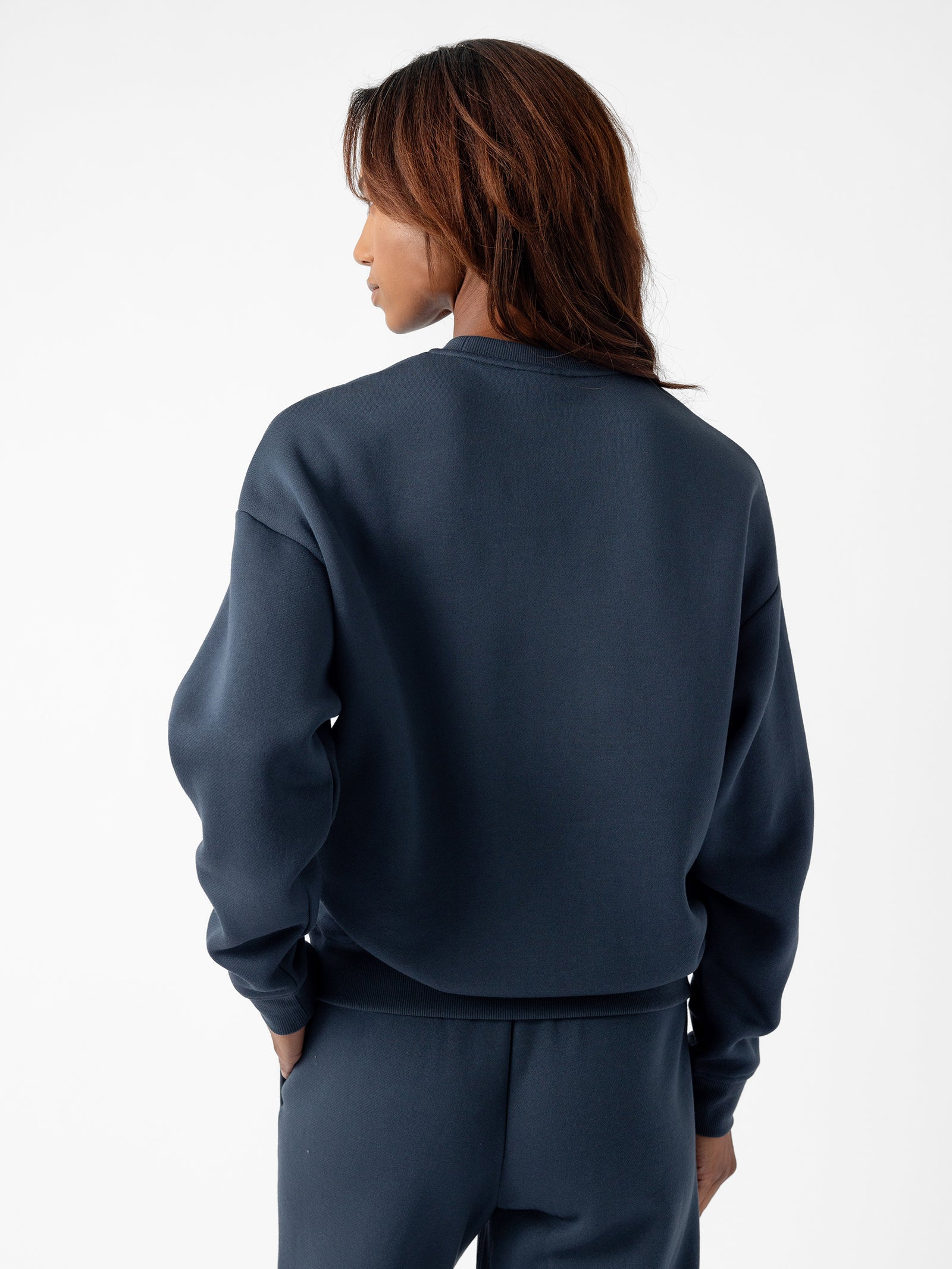 A person with shoulder-length, dark brown hair is seen from the back, wearing a dark blue Women's CityScape Crewneck by Cozy Earth and matching pants. They are standing against a plain white background. 