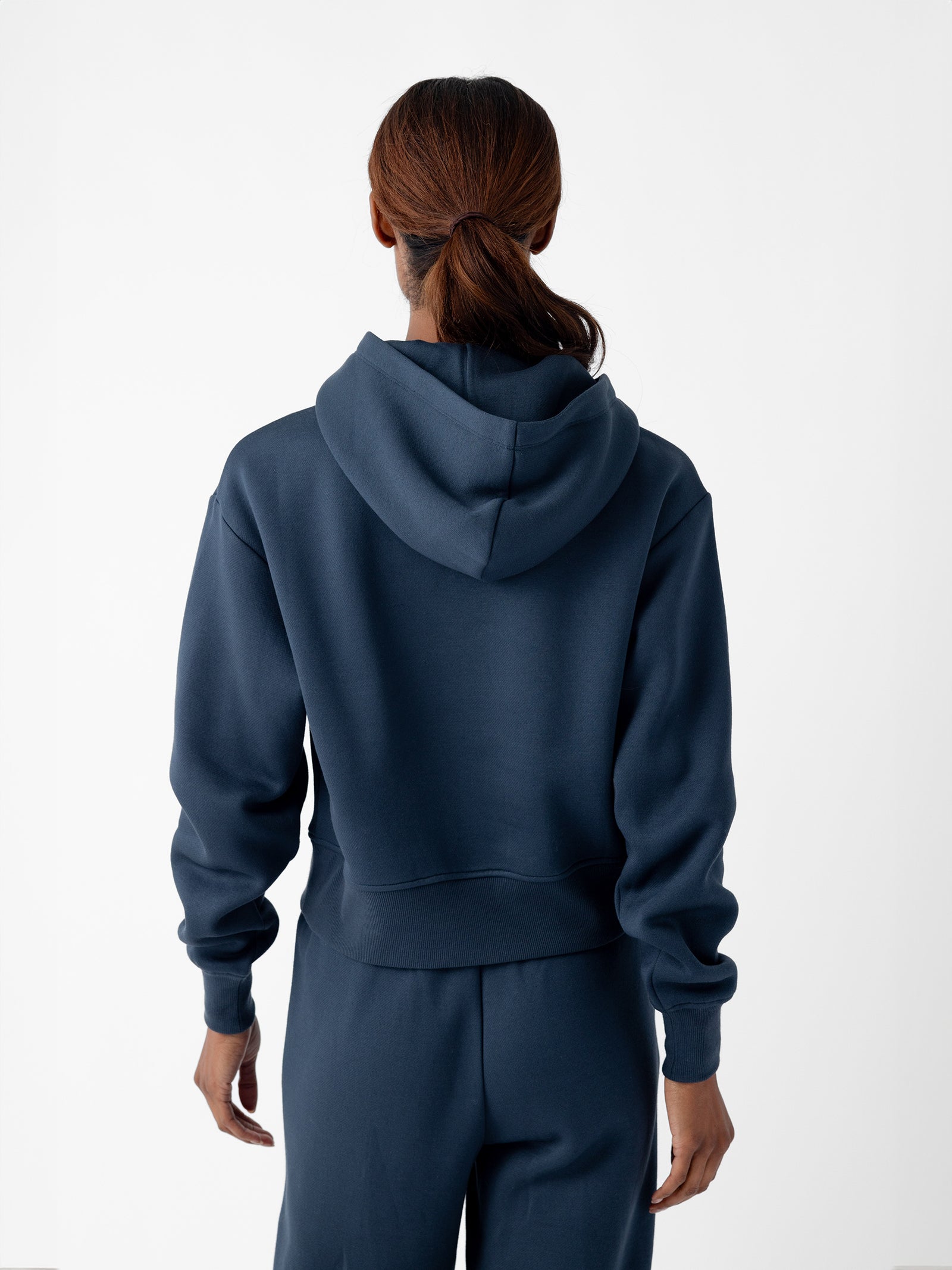 A person with long hair tied back in a ponytail is facing away from the camera. They are wearing a dark blue Women's CityScape Cropped Hoodie and matching pants by Cozy Earth. The background is plain and white. 