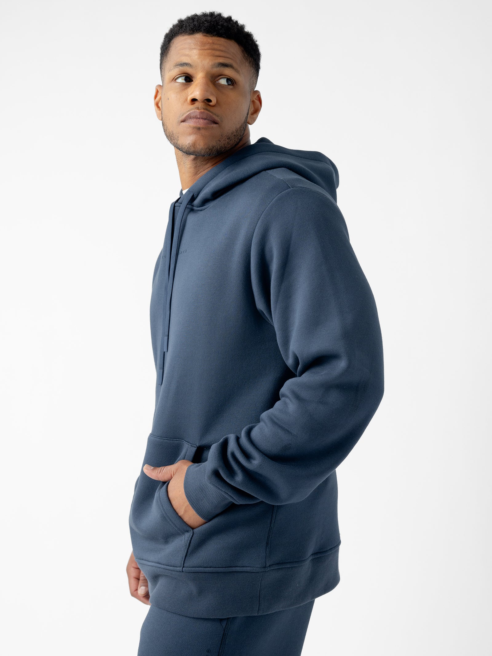 A man wearing a Cozy Earth Men's CityScape Hoodie and sweatpants stands against a plain white background. He is looking off to the side while his left hand is tucked into the hoodie pocket. The dark blue hoodie has a relaxed fit and a drawstring hood. 