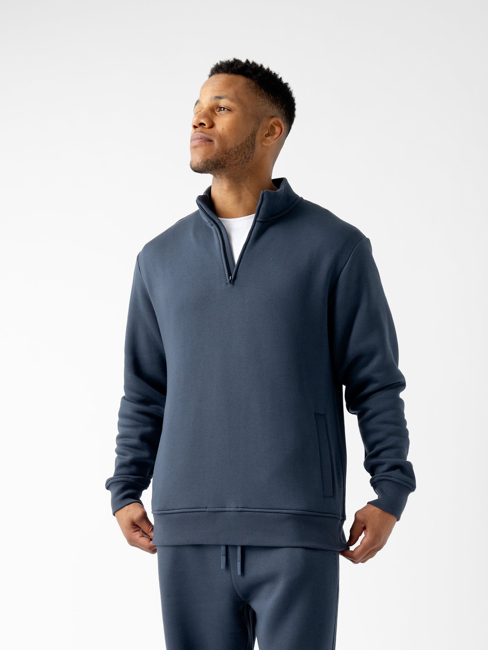 A person wearing a dark blue Men's CityScape Quarter Zip sweater by Cozy Earth over a white shirt stands against a plain white background. The individual has short hair and is looking slightly upwards and to the side with a neutral expression. 