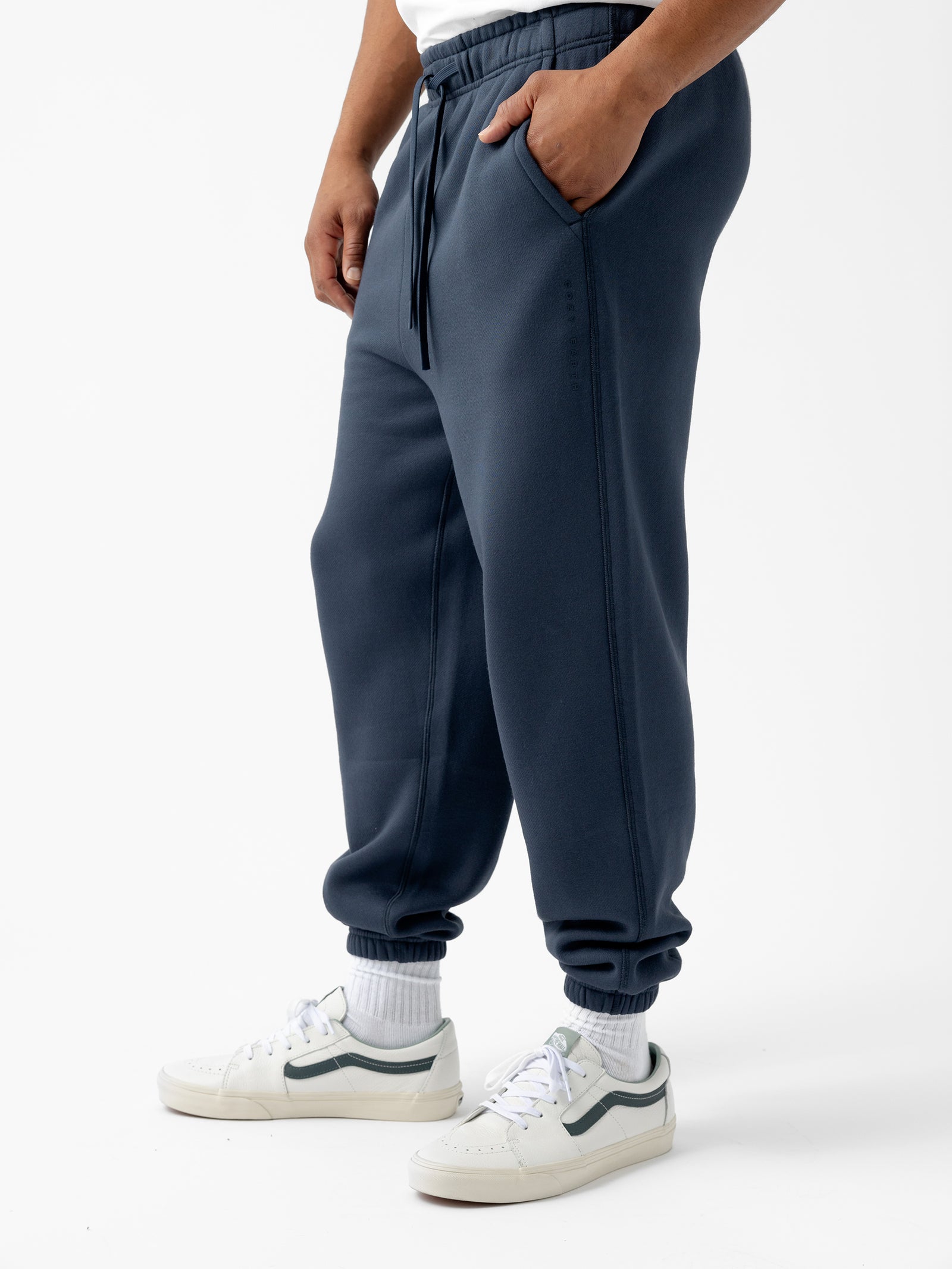 A person wearing Cozy Earth's Men's CityScape Sweatpant in dark blue with pockets, a white T-shirt, white socks, and white sneakers with dark accents stands against a plain white background. The person's hands are in the pockets of the sweatpants. 