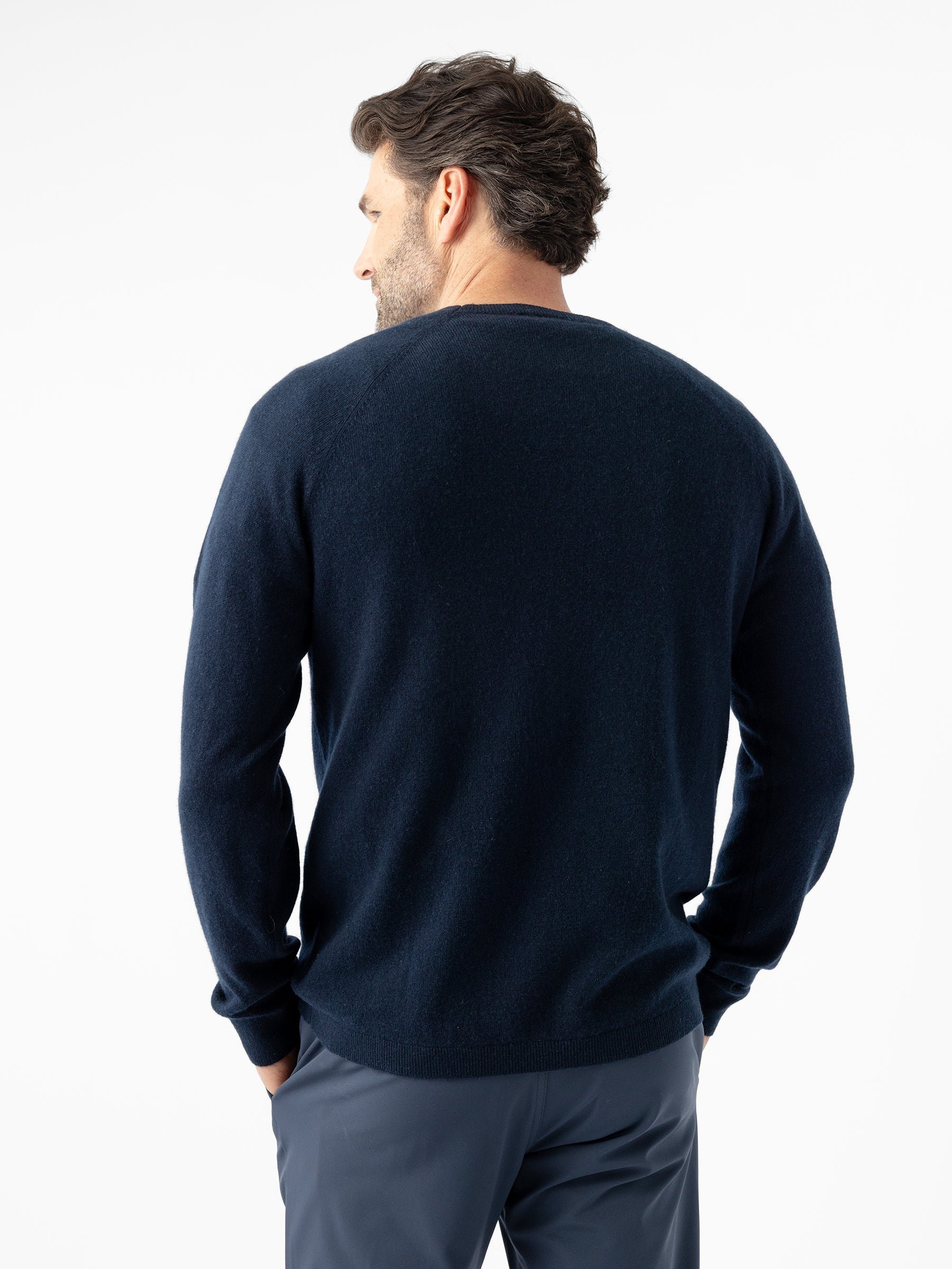 A man with dark hair and a beard, seen from behind, wears a navy blue Men's Crewneck Sweater by Cozy Earth and gray pants. He stands against a white background with his hands in his pockets. 