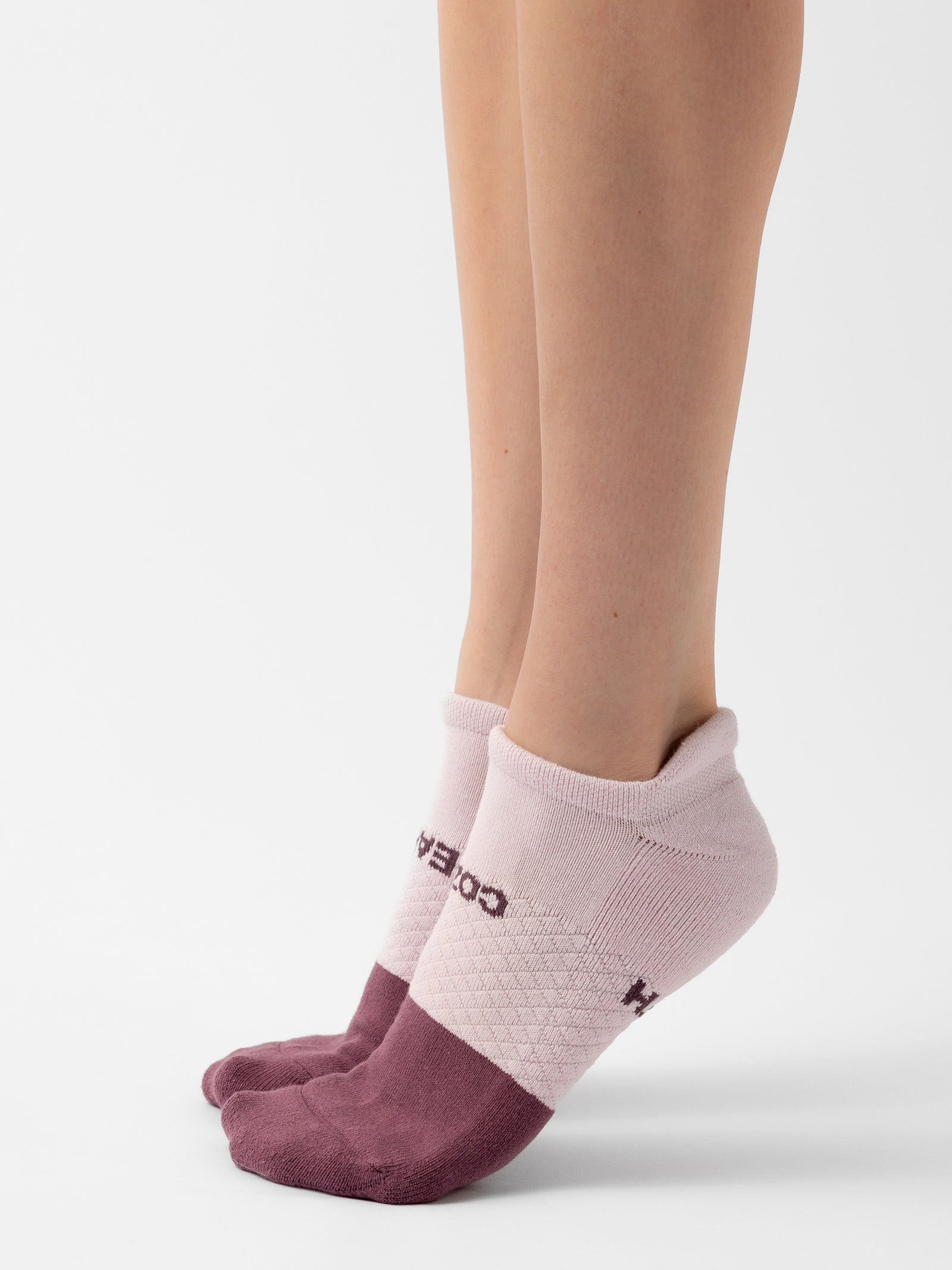 Close-up of a person's lower legs and feet wearing pink and maroon ankle socks. The socks have a textured design on the top, with the brand name "Cozy Earth" visible near the ankles. The person is standing on a white background, showcasing the Essential Ankle Sock 2-Pack. 