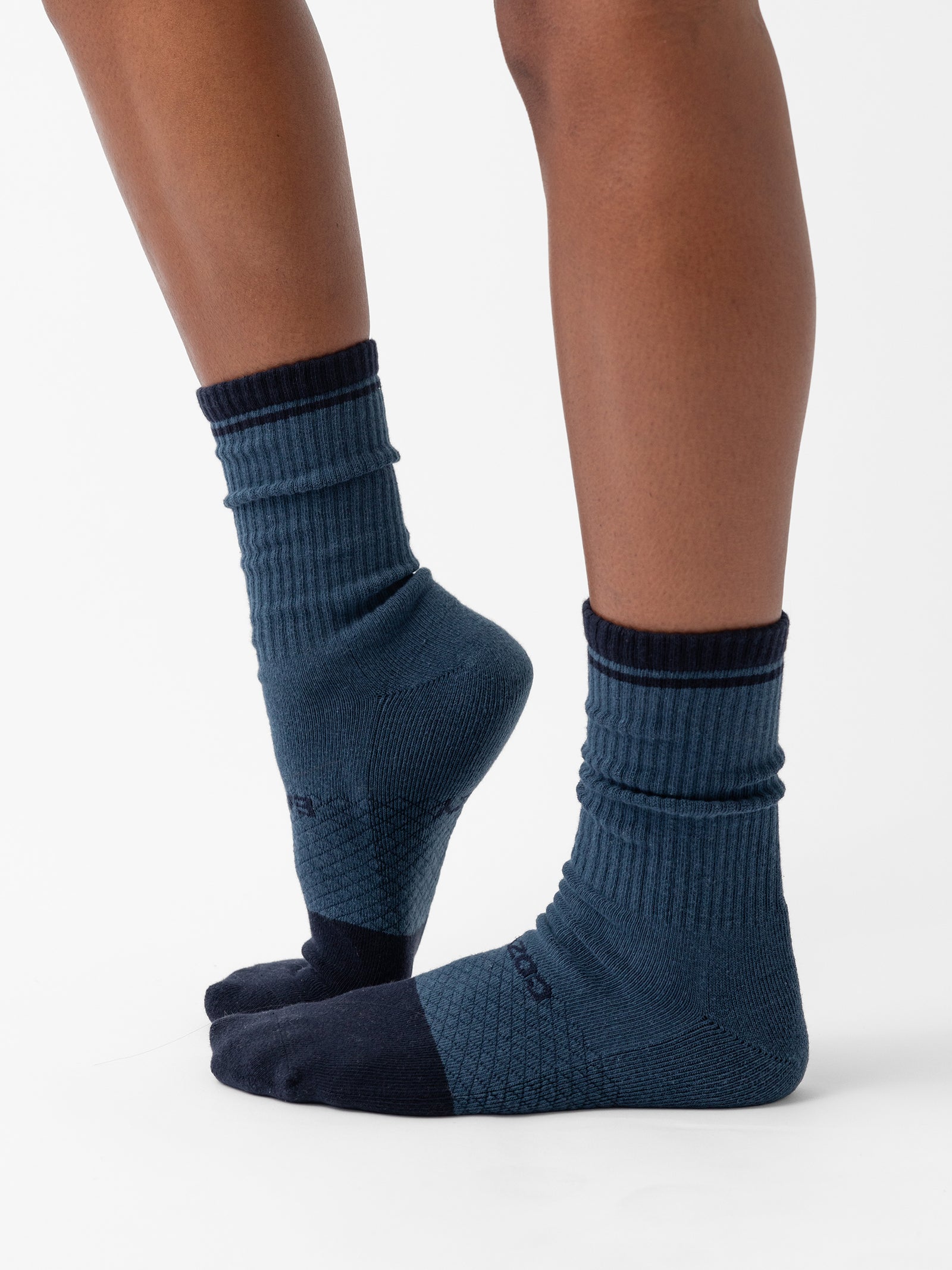 A person wearing dark blue Essential Calf Socks from the Cozy Earth 2-Pack, featuring black toes and heels, stands with one foot slightly in front of the other against a plain white background. 