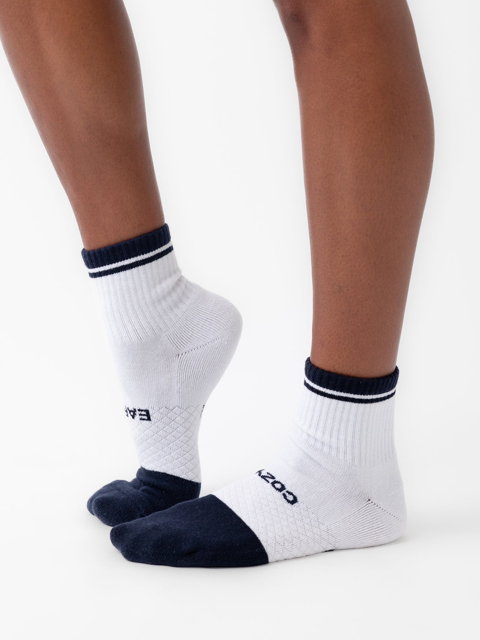 A person is standing on tiptoes, wearing a pair of Cozy Earth's Essential Quarter Socks in white, featuring black toe caps, heels, and cuffs. The socks are designed with black stripes around the cuffs and have a logo printed near the toes. The background is plain white. 