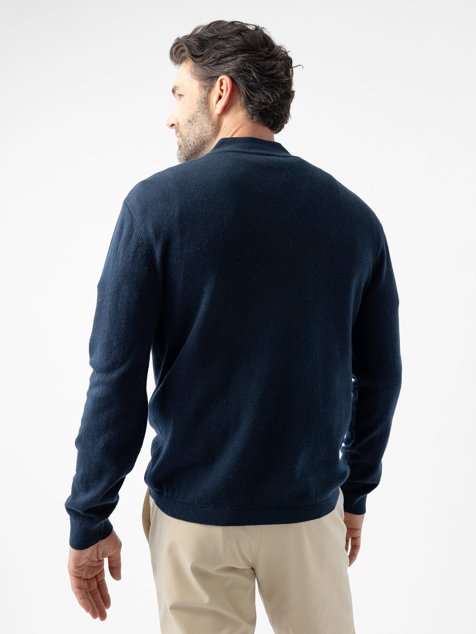 A man with dark hair and a beard is facing away from the camera, wearing a Cozy Earth Men's Quarter Zip Sweater in dark navy-blue and beige pants. The background is plain white. 