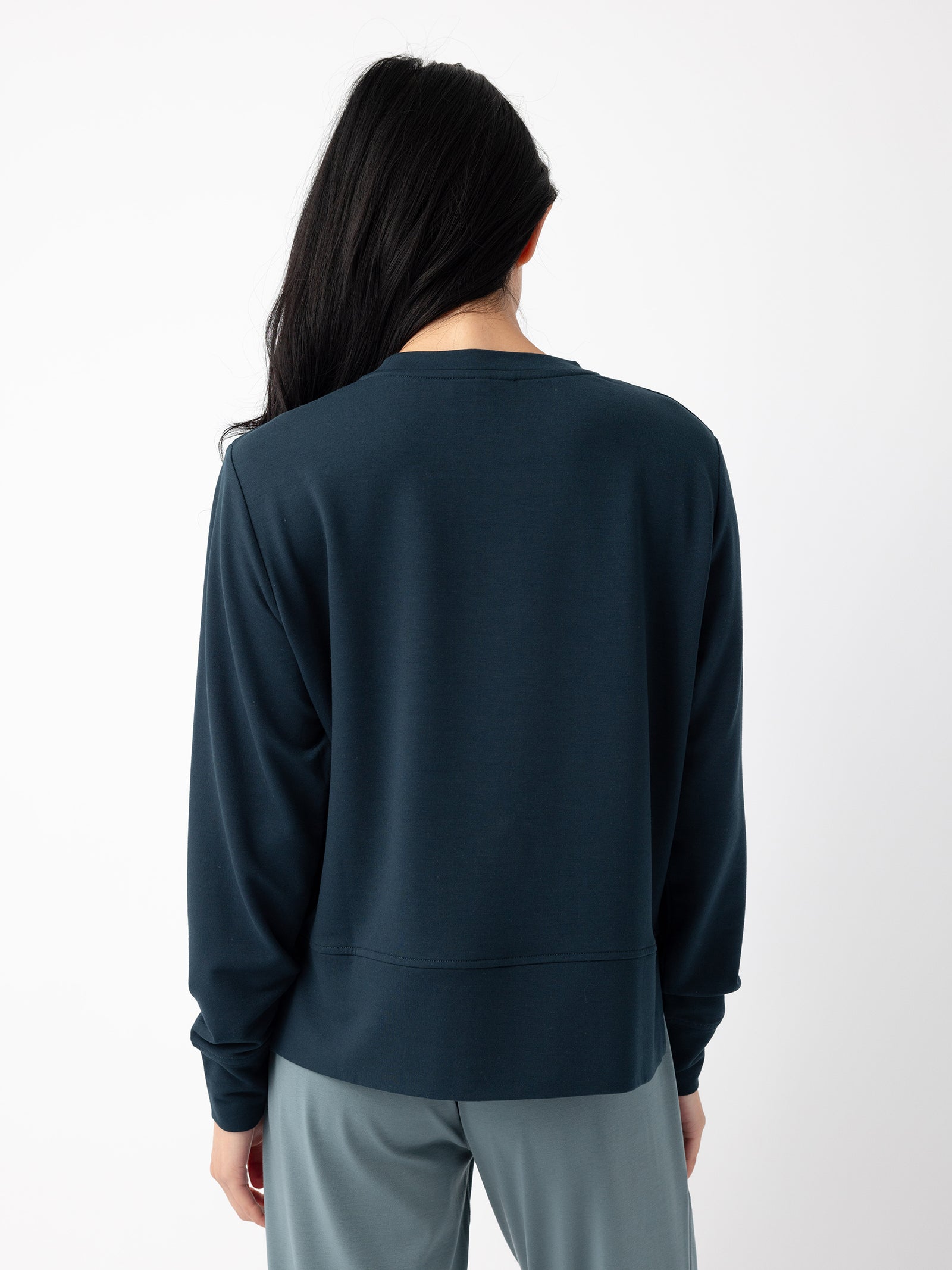 A person with long, dark hair is standing with their back to the camera, wearing a dark blue Women's StudioLite Crewneck from Cozy Earth and light blue pants against a plain white background. 