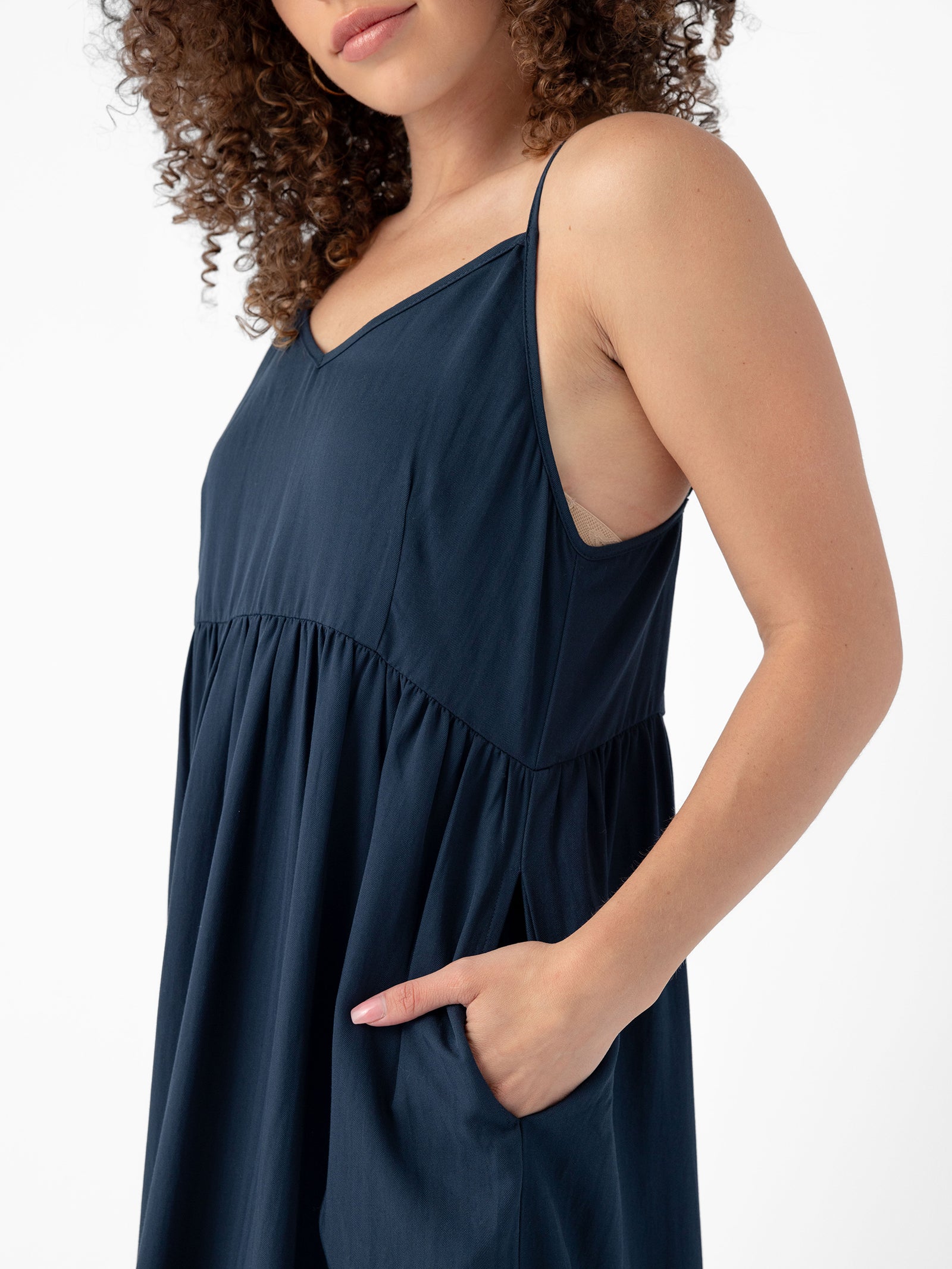 A person with curly hair is wearing a Women's Sunset Gathered Dress by Cozy Earth. The sleeveless, dark blue dress features a V-neckline and an empire waist. The person is casually placing a hand in one of the dress's side pockets against a plain white backdrop. 