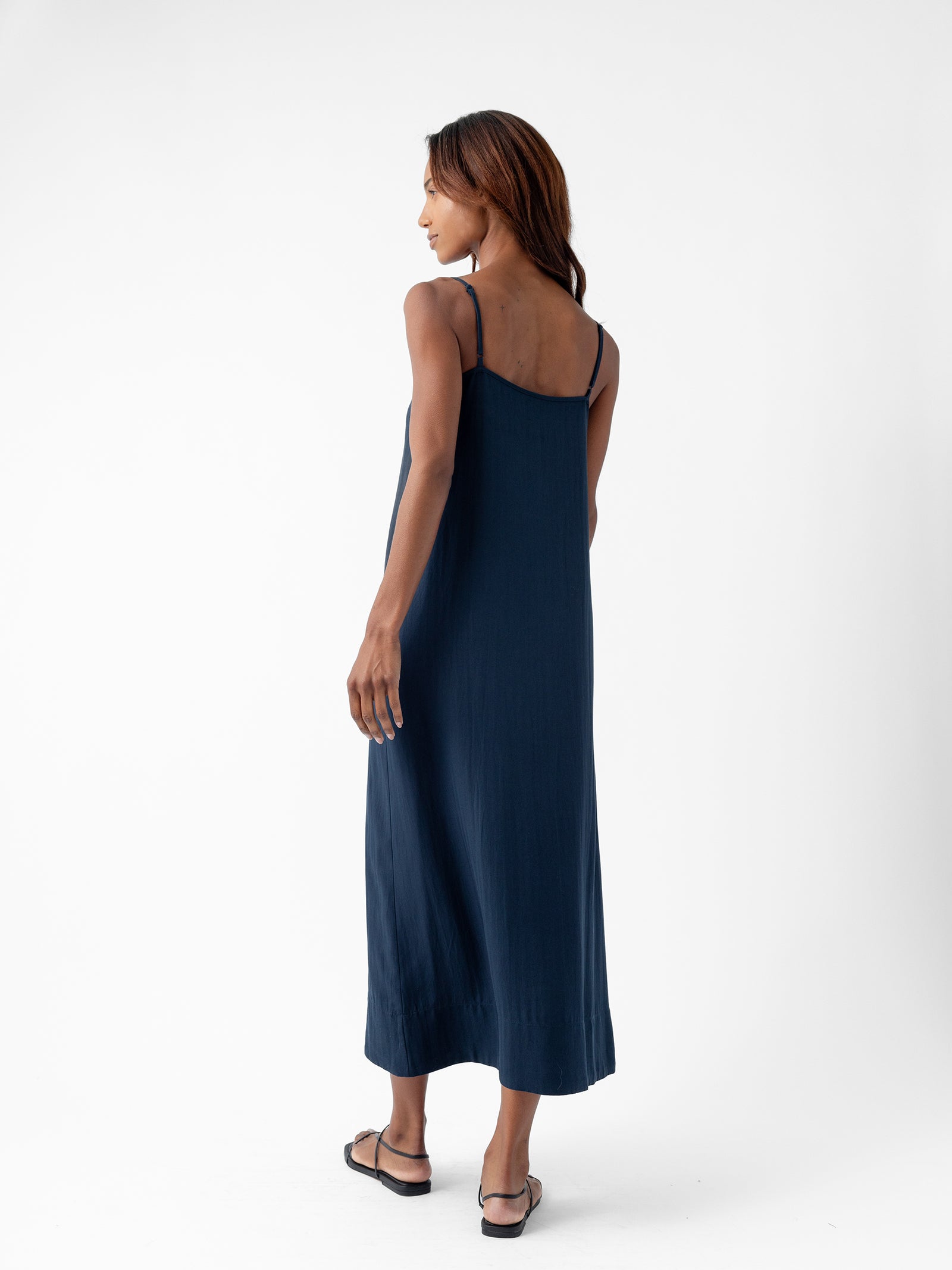 A woman with long hair is standing against a plain white background. She is wearing the Women's Sunset Slip Dress in navy blue from Cozy Earth and black flip-flops. The woman is turned slightly to the side, looking towards the right, showcasing the back of her dress. 