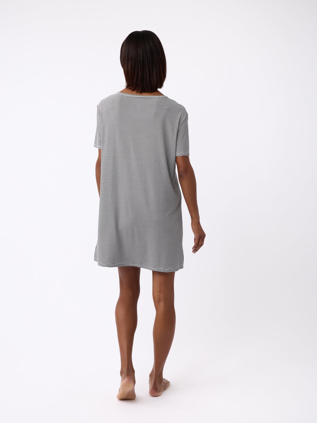 A person with short dark hair stands barefoot on a white background, wearing a loose-fitting gray Women's Bamboo Stretch-Knit Sleep Dress by Cozy Earth, viewed from the back. 