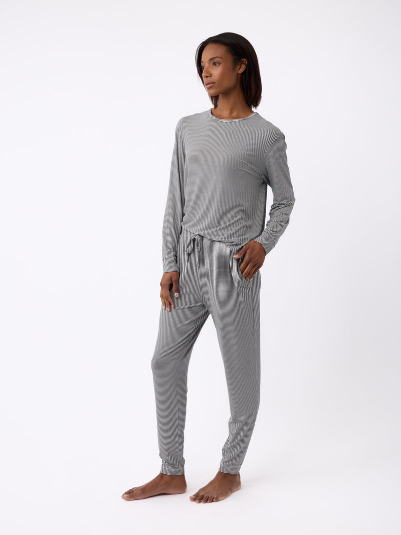 A person stands barefoot against a white background, wearing Cozy Earth's Women's Bamboo Stretch-Knit Long Sleeve Lounge Tee in gray with matching pants. With one hand in their pocket, they exude calmness and relaxation.