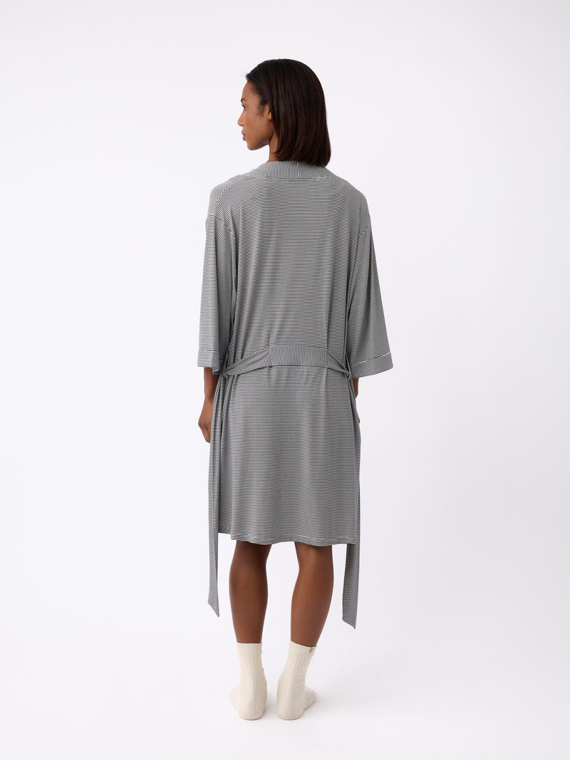 A person stands facing away in a Cozy Earth Women's Bamboo Stretch-Knit Kimono Robe, featuring loose sleeves and a waist tie, paired with light-colored socks against a white background.