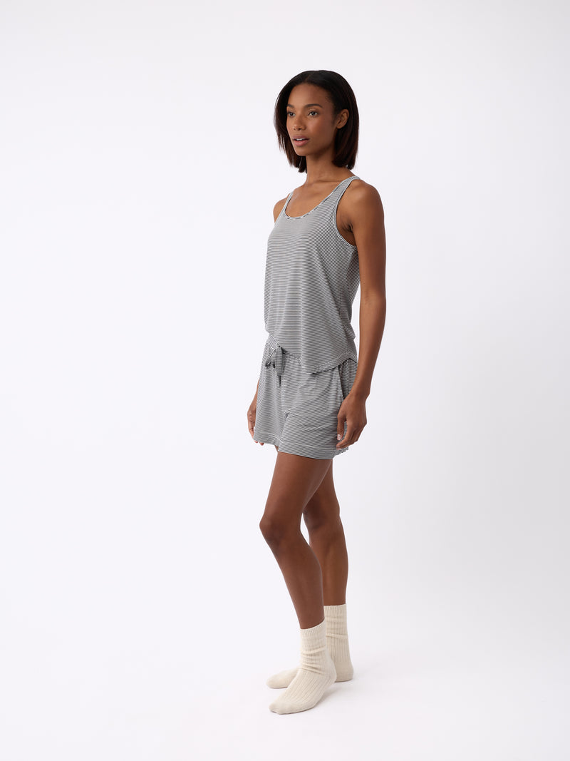 A person with short dark hair stands left-facing against a white background, wearing Cozy Earth's light gray Women's Bamboo Stretch-Knit Shorts and a sleeveless top, paired with light-colored socks.