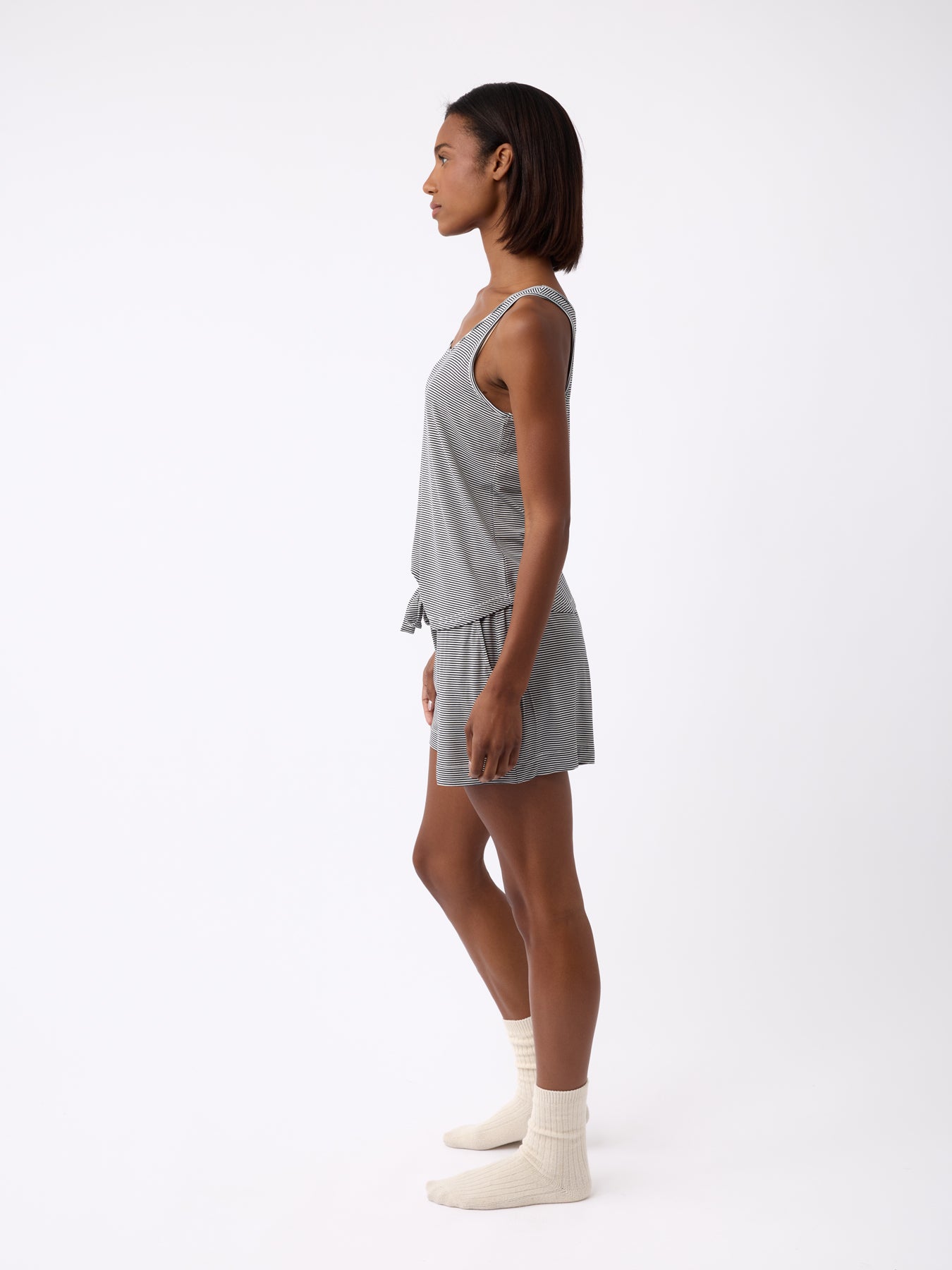 A person stands in profile against a plain white background, wearing the Women's Bamboo Stretch-Knit Lounge Tank by Cozy Earth, paired with matching shorts and cream-colored socks. 