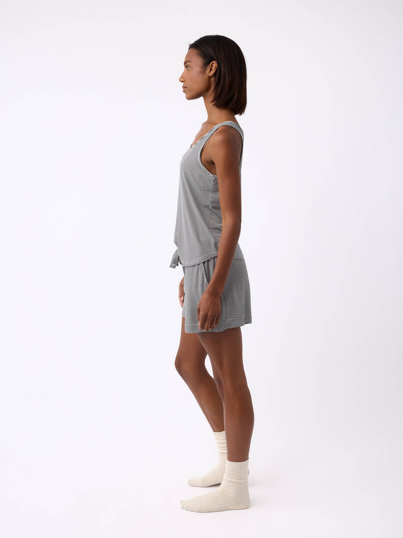A person stands in profile against a plain white background, wearing the Women's Bamboo Stretch-Knit Lounge Tank by Cozy Earth, paired with matching shorts and cream-colored socks.