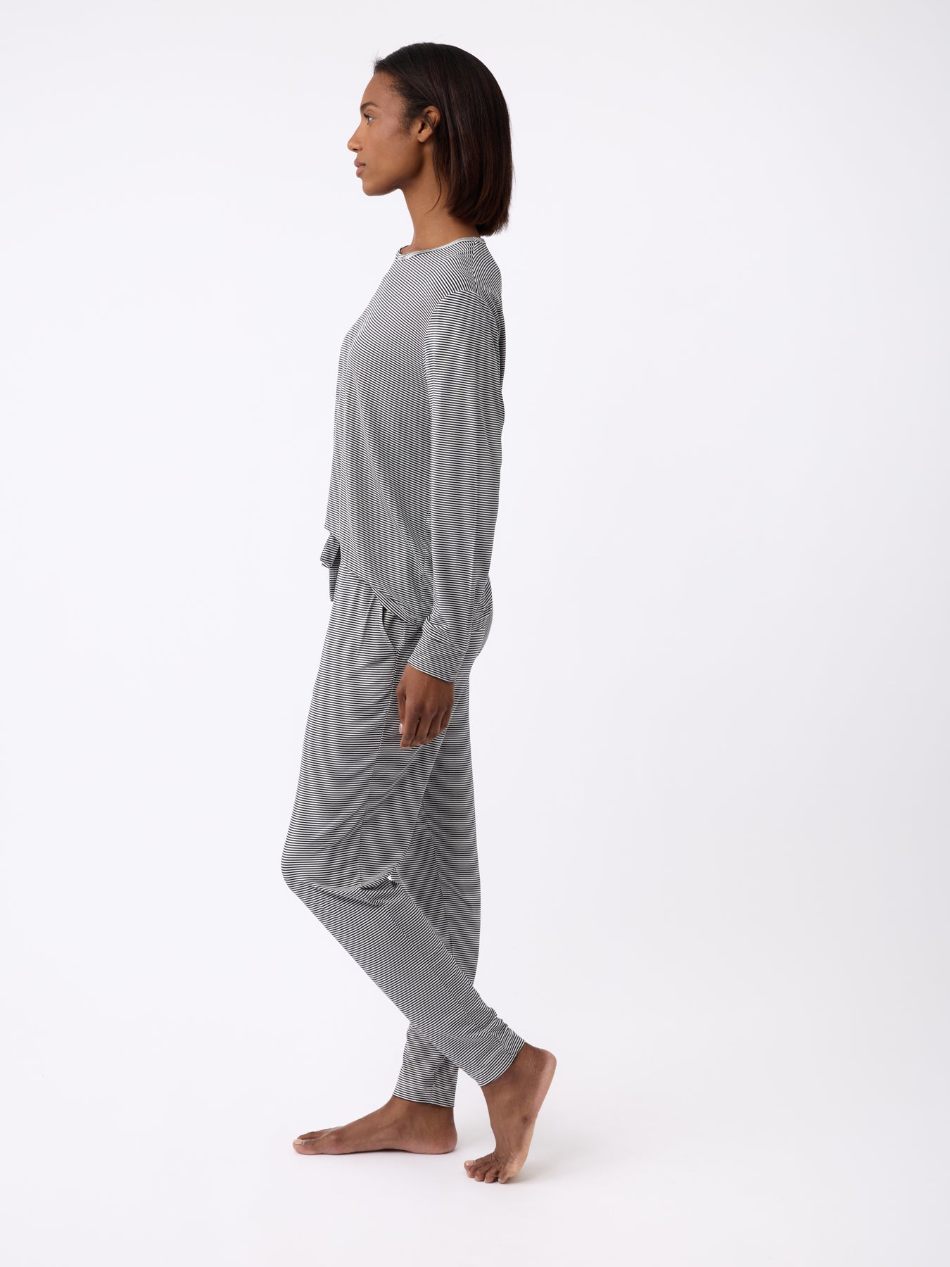 A person stands barefoot in profile, dressed in Cozy Earth's grey long-sleeve top and Women's Bamboo Stretch-Knit Tapered Pant, set against a simple white backdrop. 